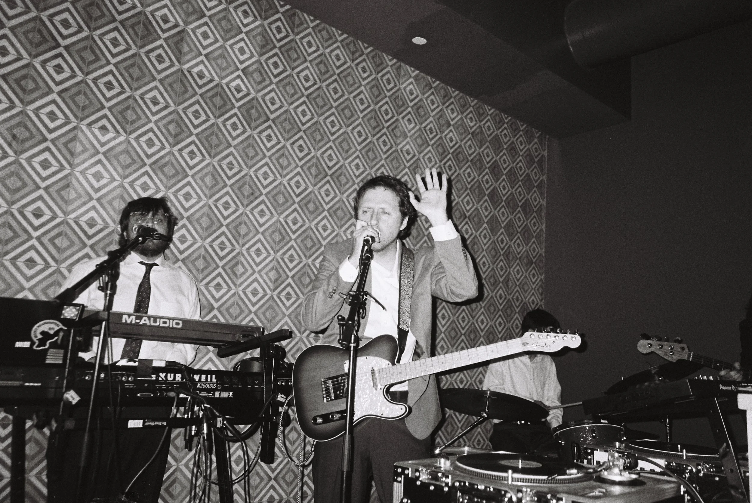 Black and white photo of three musicians performing on stage. The central figure is a man singing into a microphone while playing an electric guitar, raising his left hand. To his left, another musician is playing a keyboard with a microphone. To the right, a partially visible person appears to be playing a guitar, and there is a turntable in the foreground.
