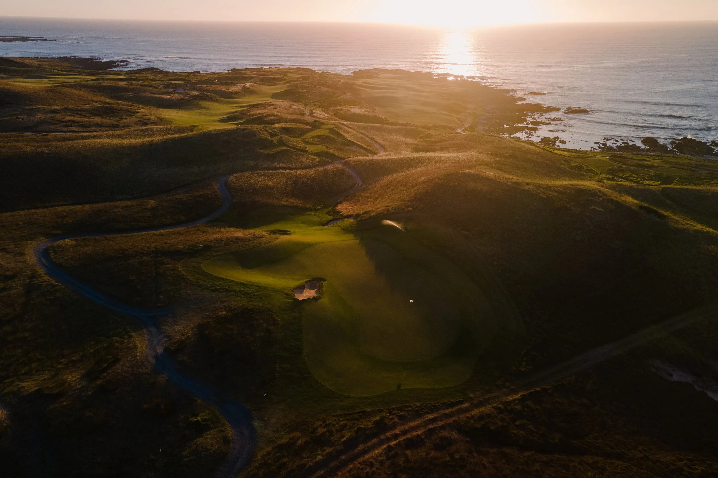 Aerial view of a golf course overlooking the ocean at sunset, with winding roads and lush green fairways.