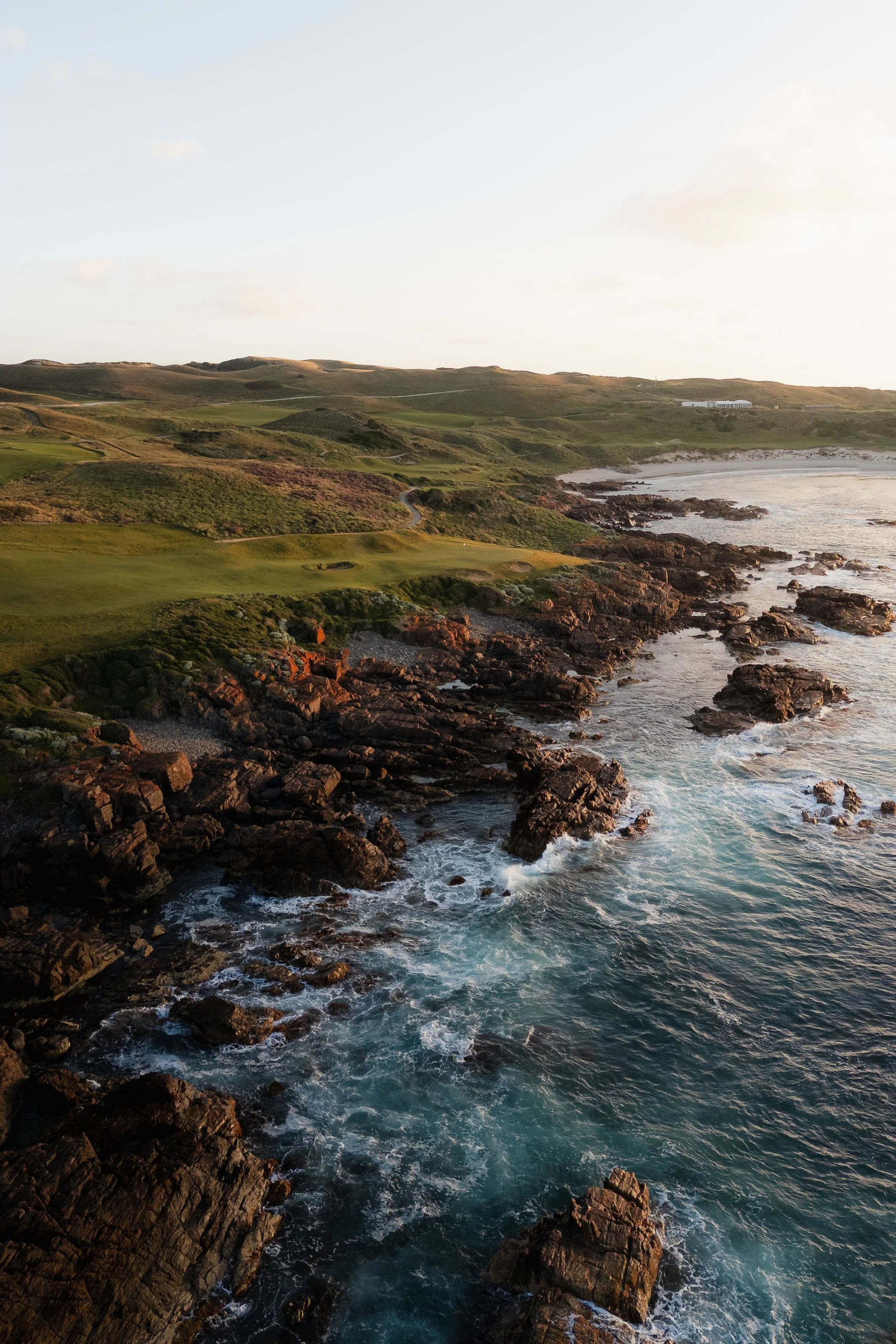 Coastal landscape with rocky shoreline, waves crashing against rocks, grassy hills, and a few buildings in the background during sunset.