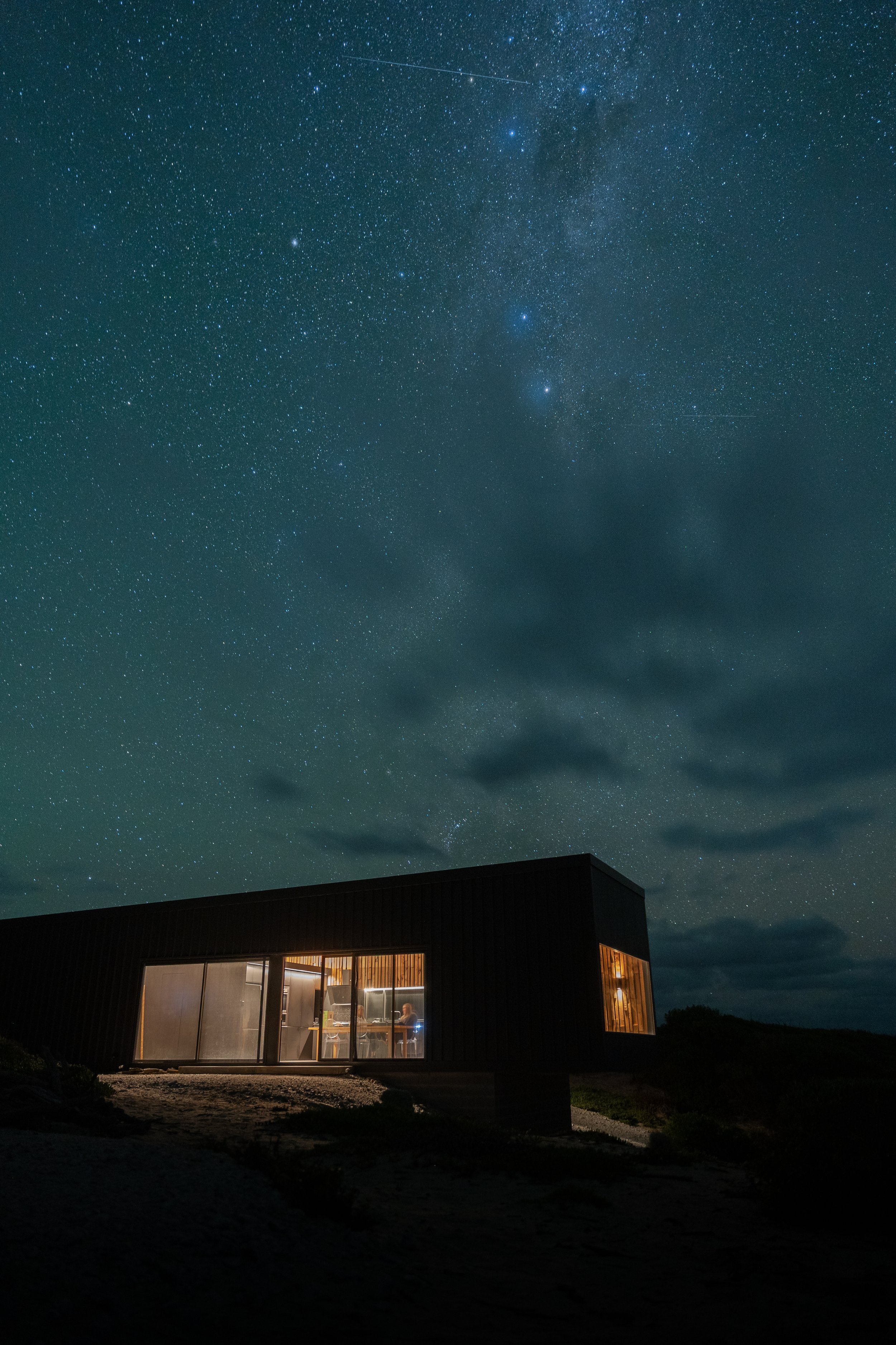 Night sky filled with stars and the Milky Way galaxy above a house with illuminated windows.