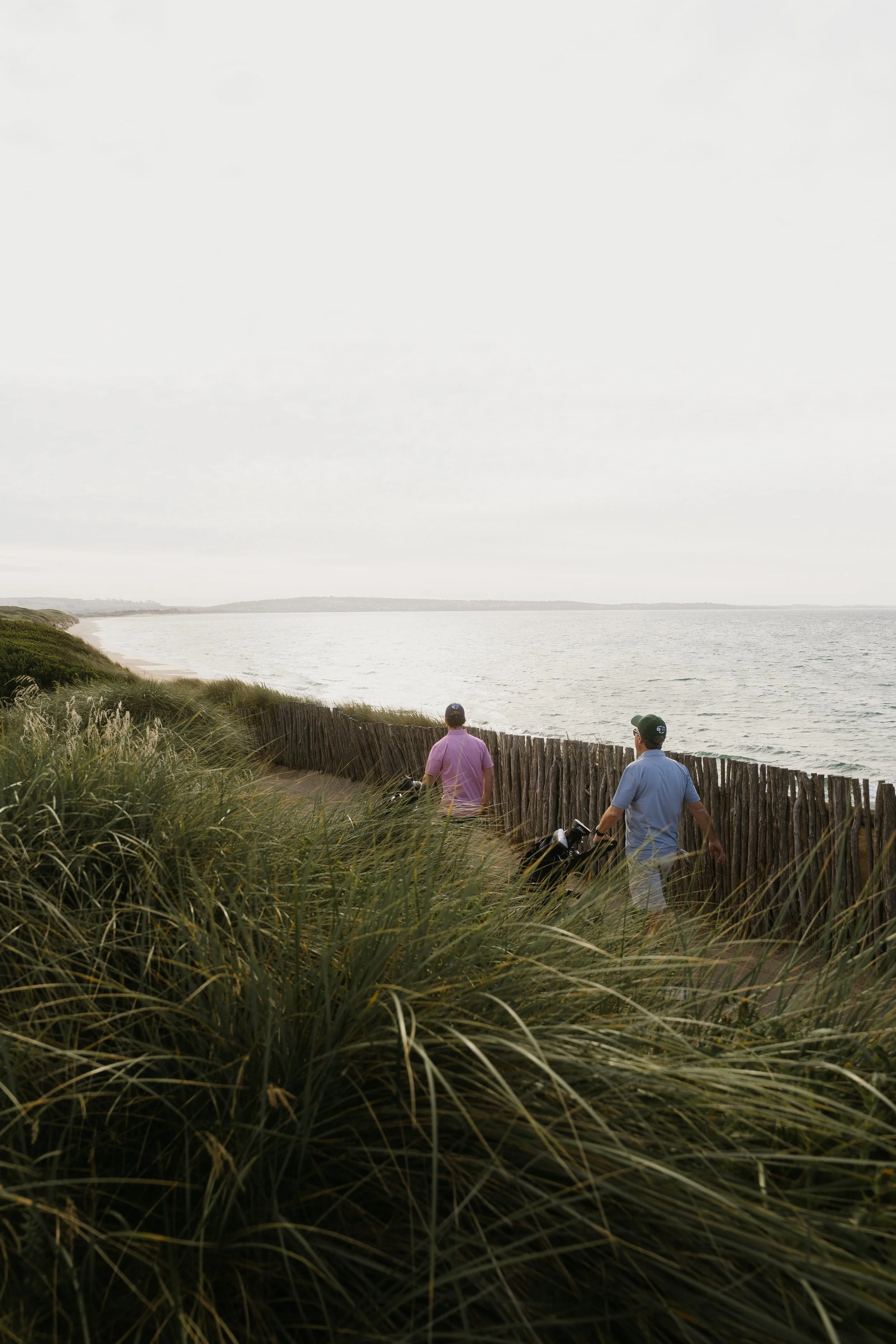Two people walking along a beachside trail with grass and a wooden fence. The ocean and cloudy sky are in the background.