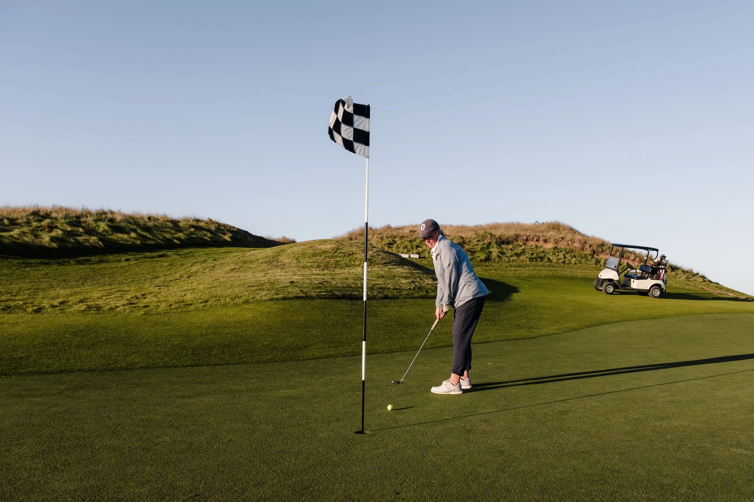 A man playing golf on a green golf course with a golf cart and a checkered flag near the hole under a clear blue sky.