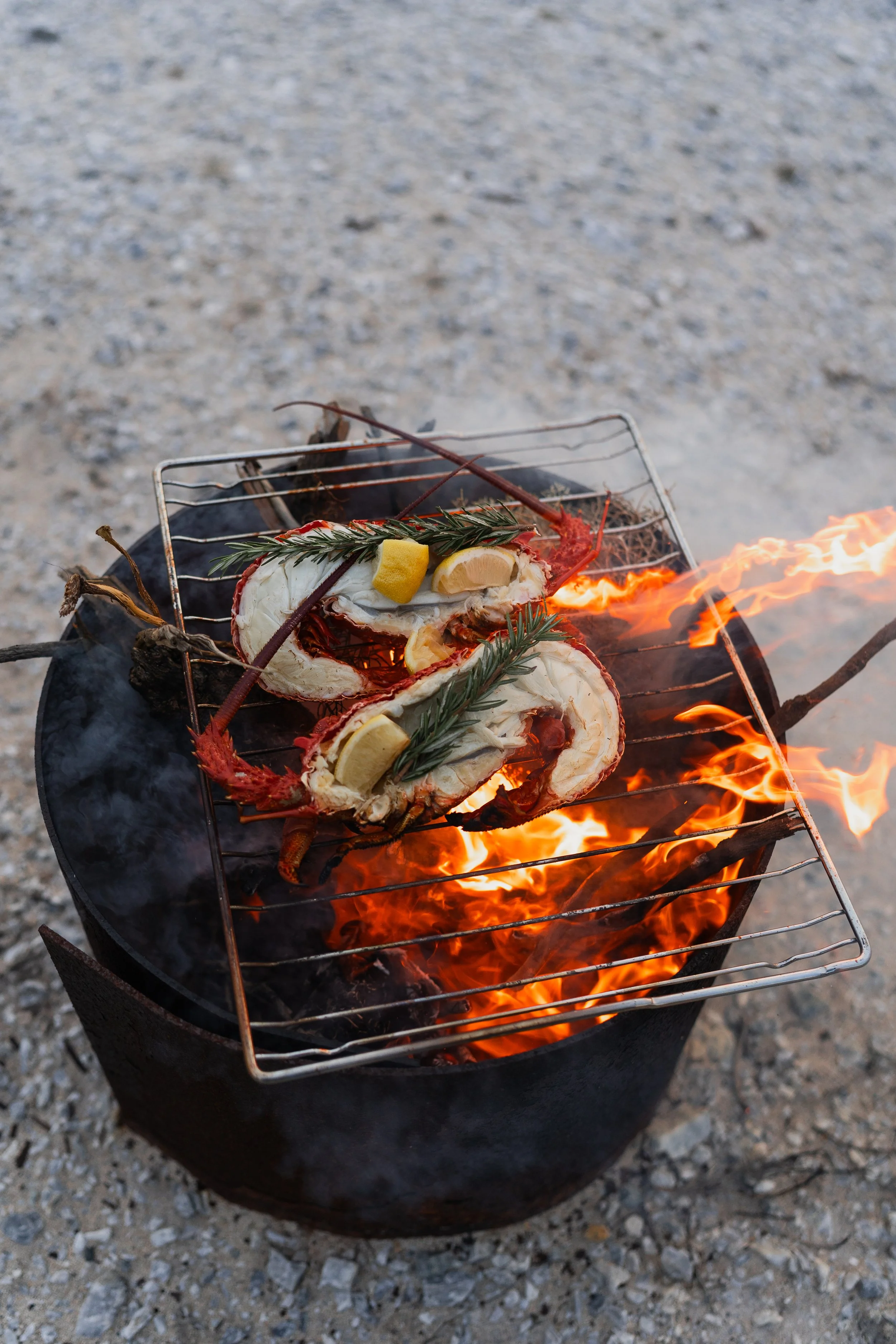 Lobster tail on a grilling rack over an open flame with herbs and lemon slices, set on a gravel surface.