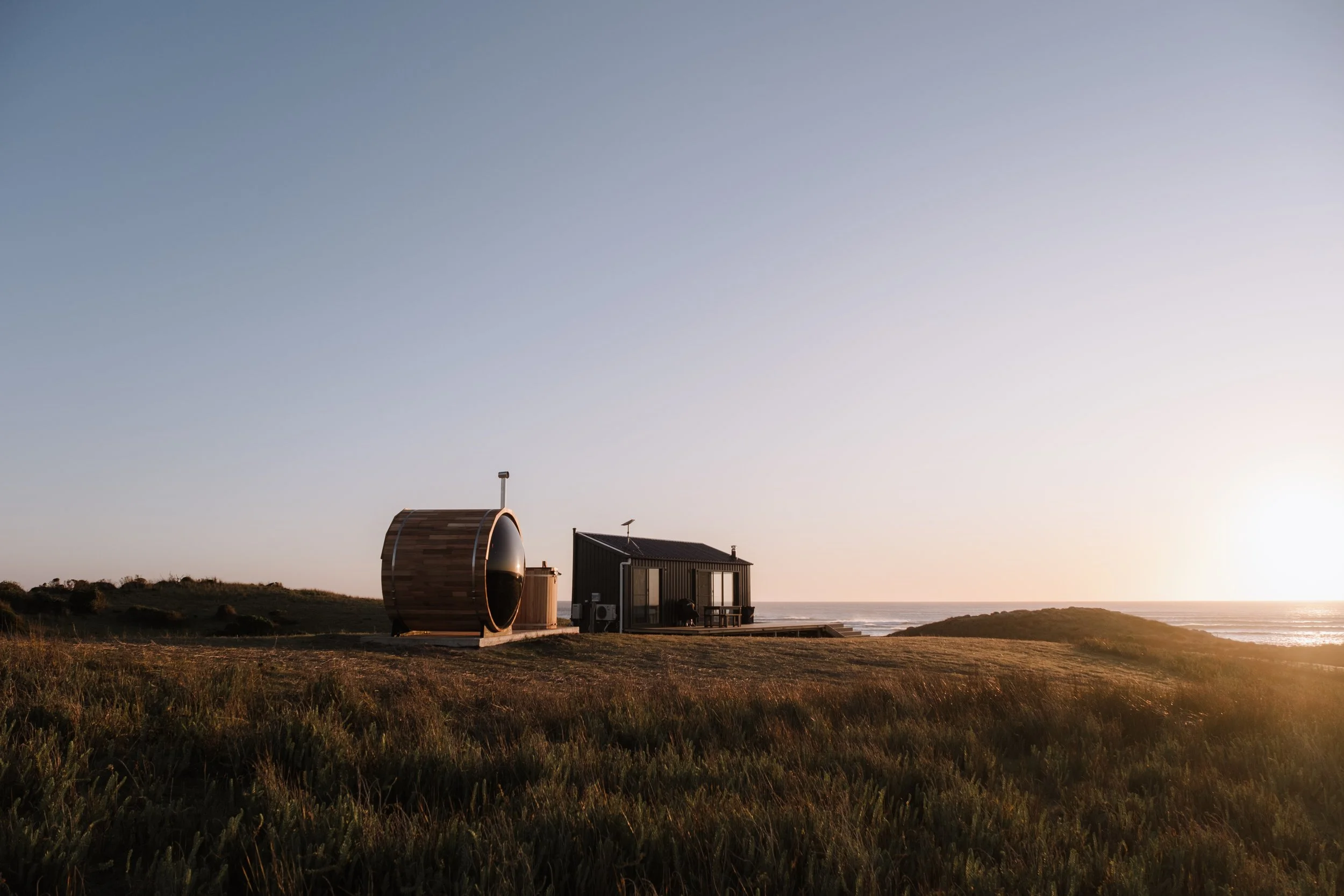 Beachfront tiny house with curved wooden sauna and modern small house, grassy landscape at sunset.