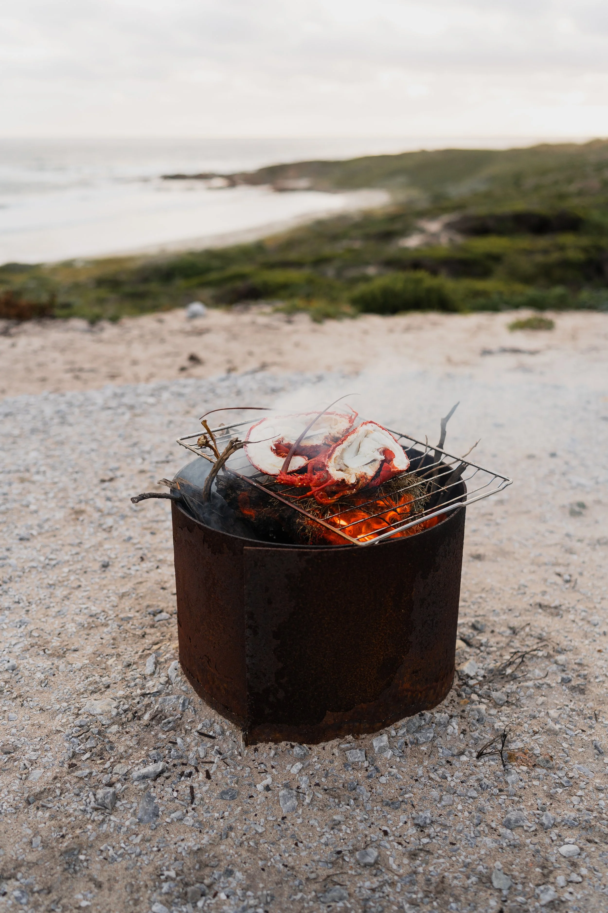 Small barbecue grill with a lobster tail on top, situated on sandy ground with rocks, beach, and ocean in the background.