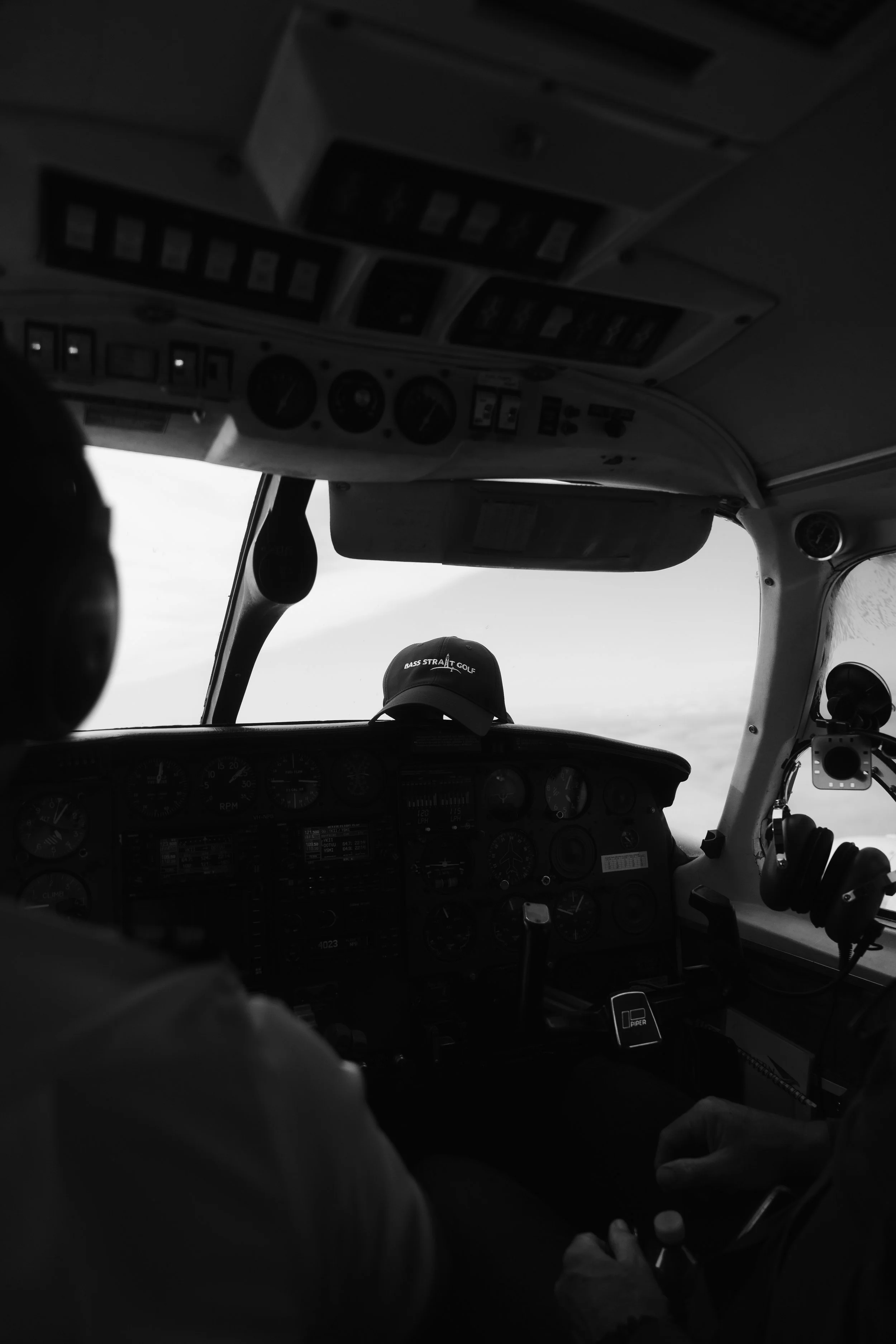 Black and white photograph of the cockpit of an aircraft, showing various instruments and controls on the dashboard, with a pilot holding a throttle or other control in their hand, and a baseball cap on the dashboard with the words "BASS STRAIT GOLF"