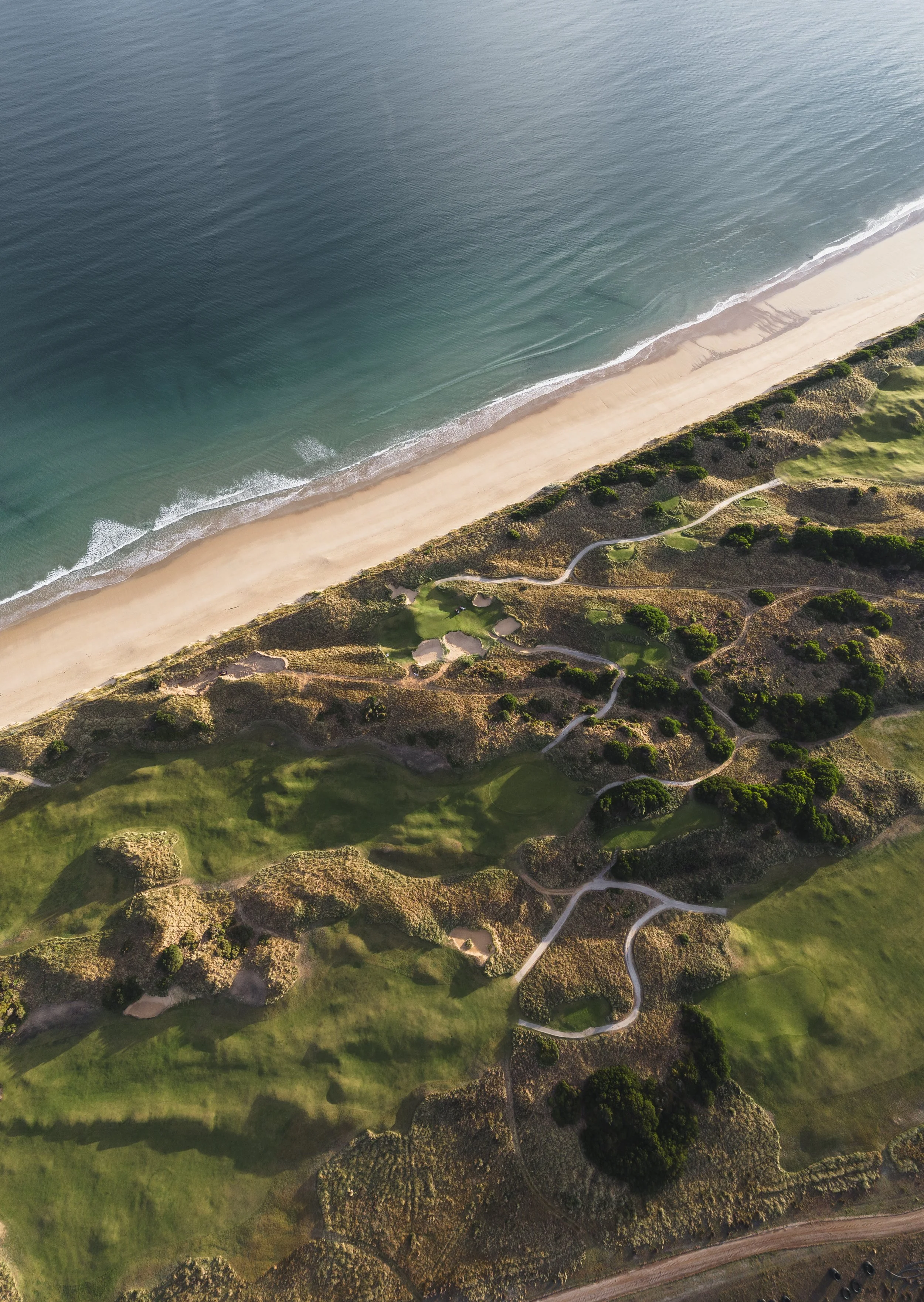 An aerial view of a beach with waves crashing onto the sandy shore, adjacent to a golf course with trees and winding pathways.