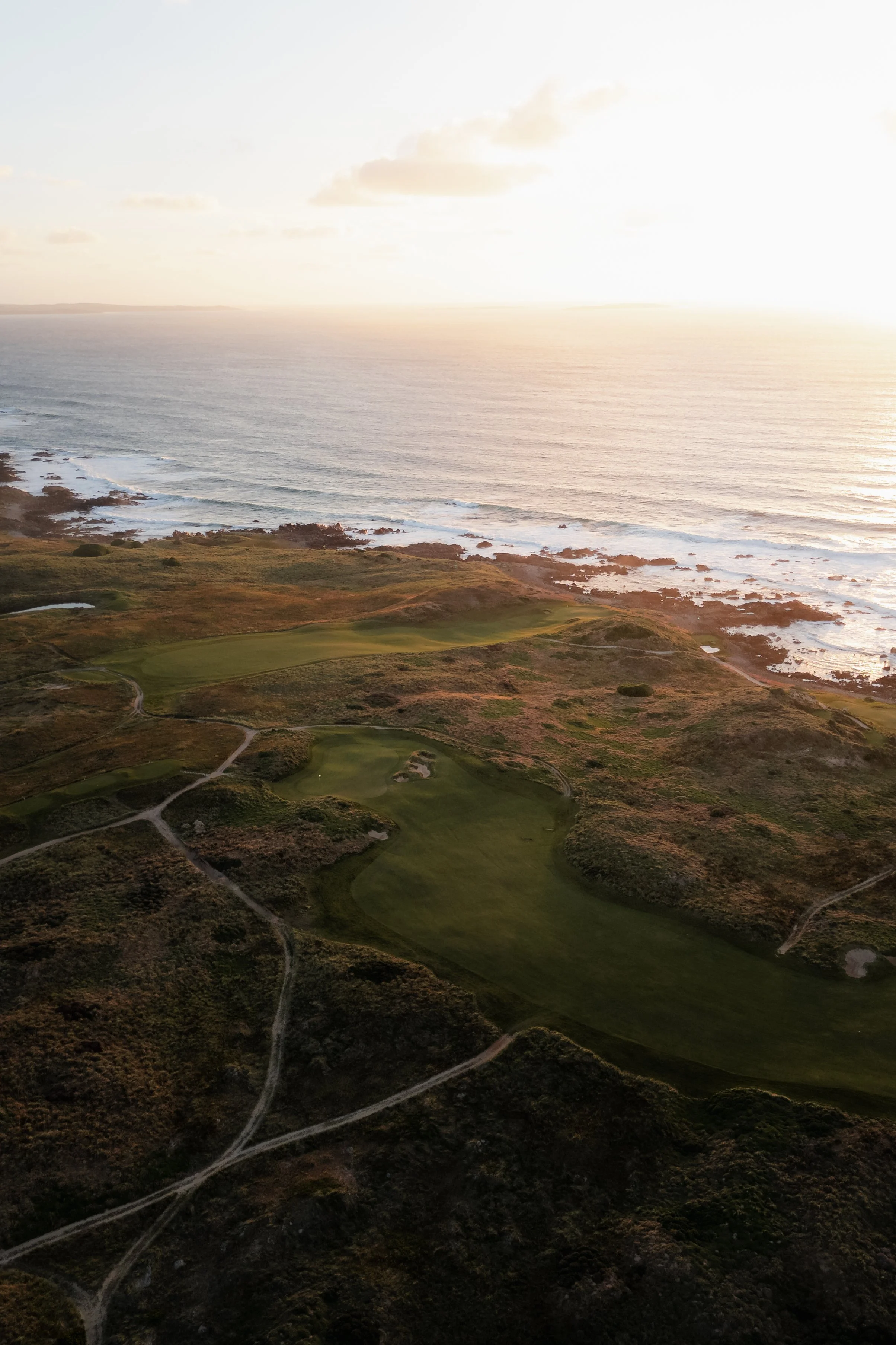 A coastal landscape showing a golf course on rolling green terrain, with paths and bushes, against the backdrop of the ocean and a setting sun, with some clouds in the sky.