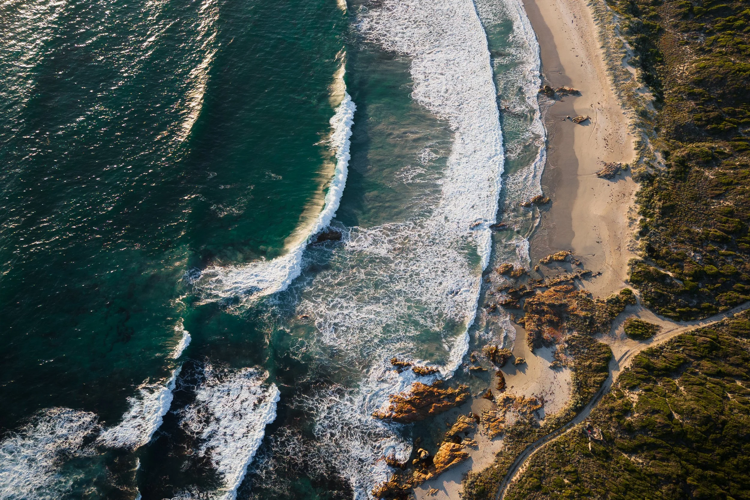 An aerial view of a rocky coastline with sandy beach and green vegetation, with waves crashing onto the shore.