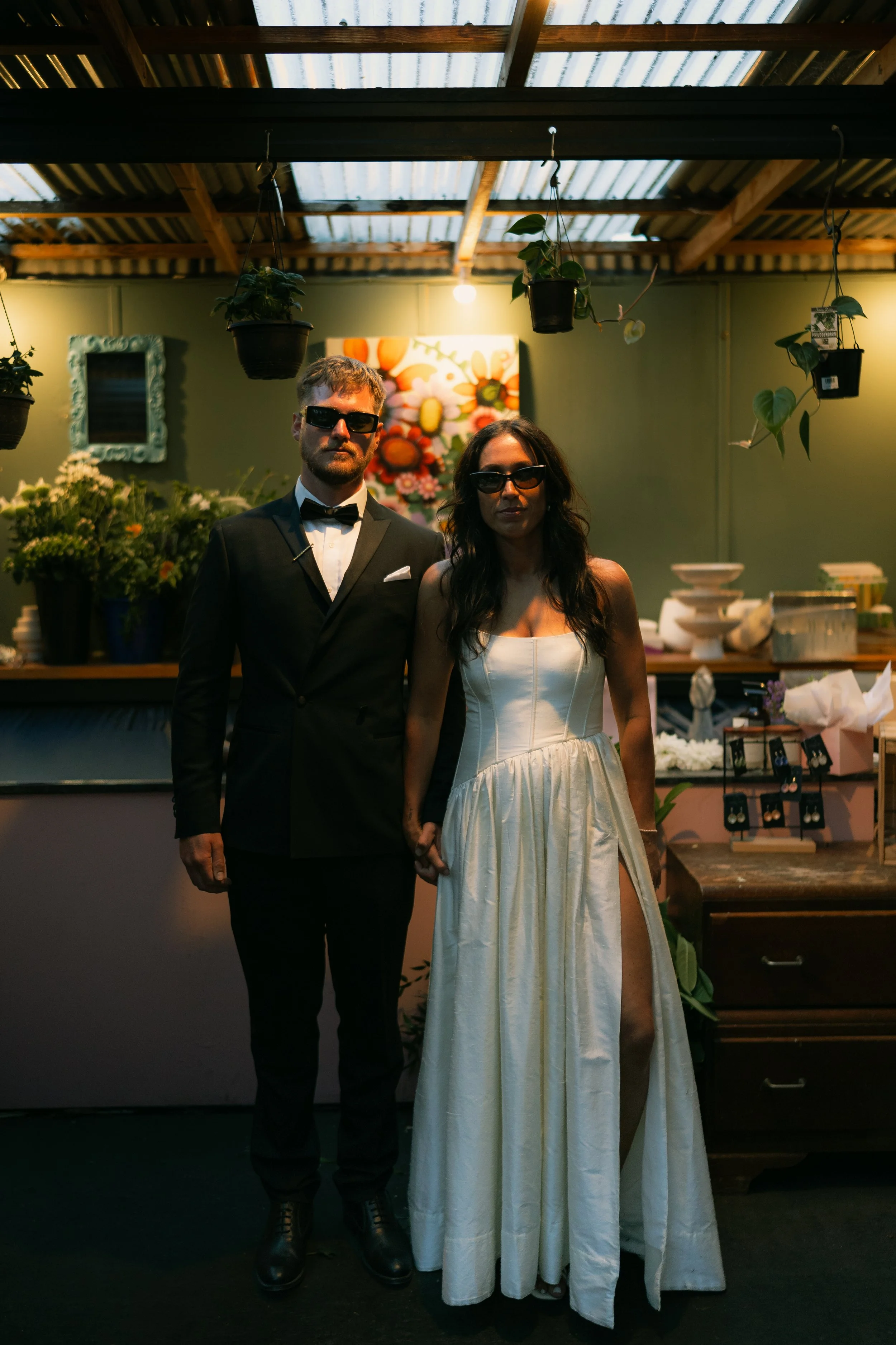 A man in a tuxedo with a bow tie and sunglasses standing next to a woman in a white dress and sunglasses, holding hands. They are inside a room with wooden ceiling beams, hanging potted plants, and decorated with artwork and plants in the background.
