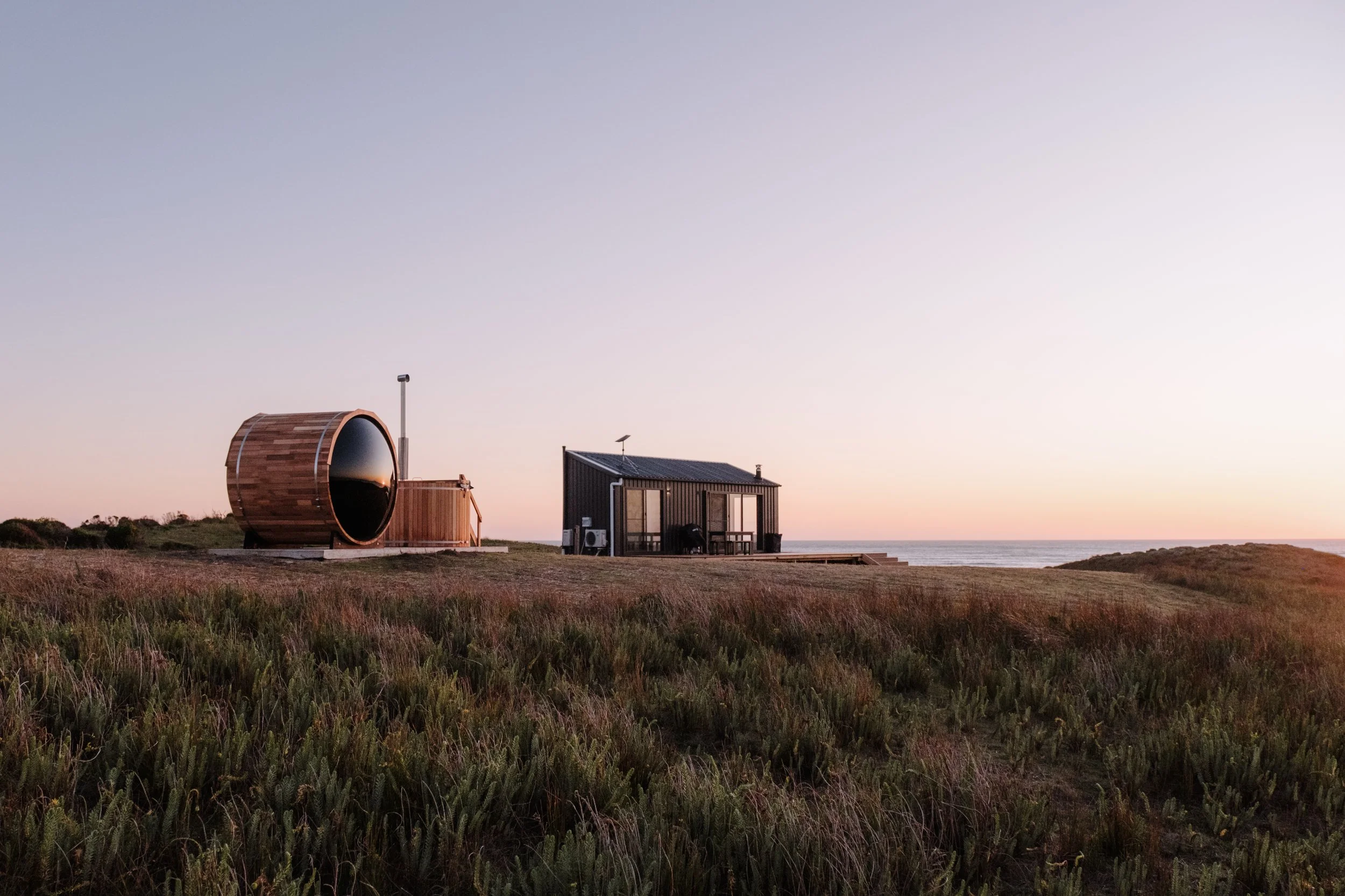 A small house and a modern tiny cabin or sauna on a grassy field near the ocean at sunset.