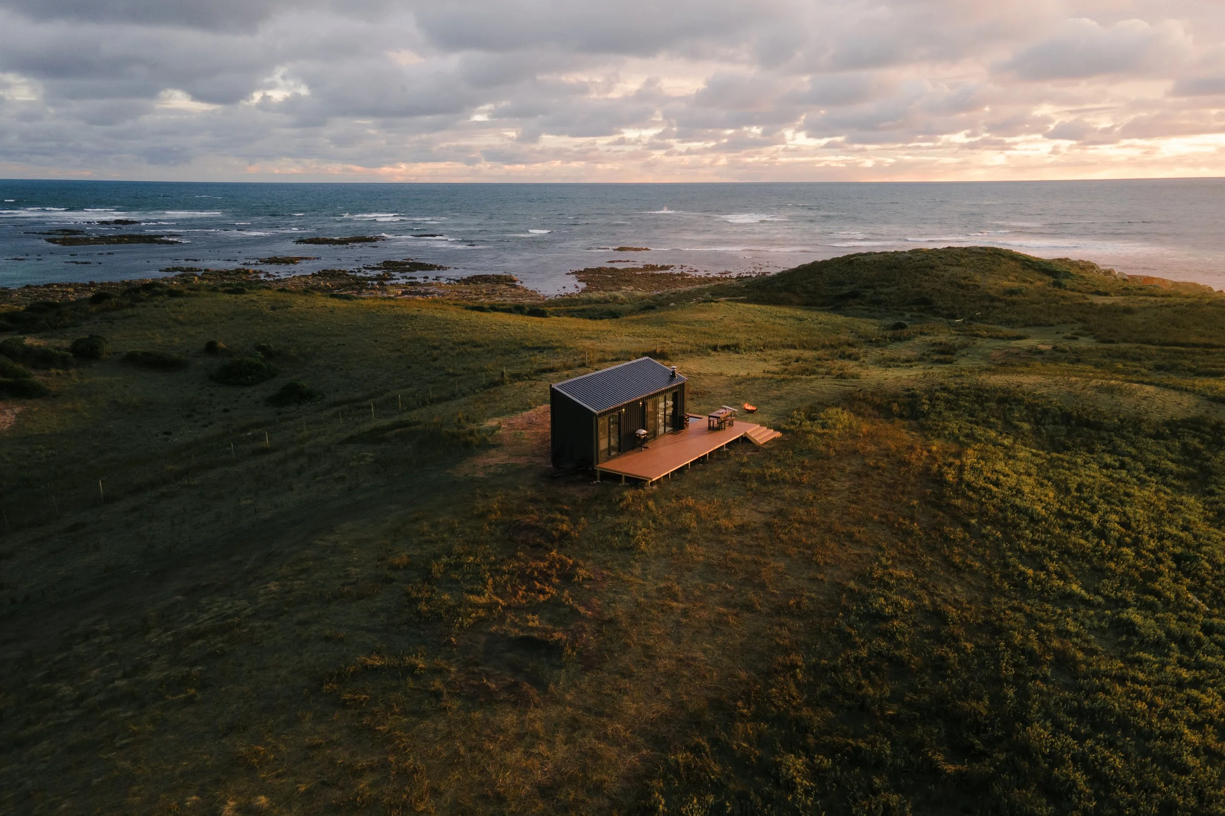 A small modern house on a grassy hill overlooking the ocean with rocky shores, under a partly cloudy sky during sunset.