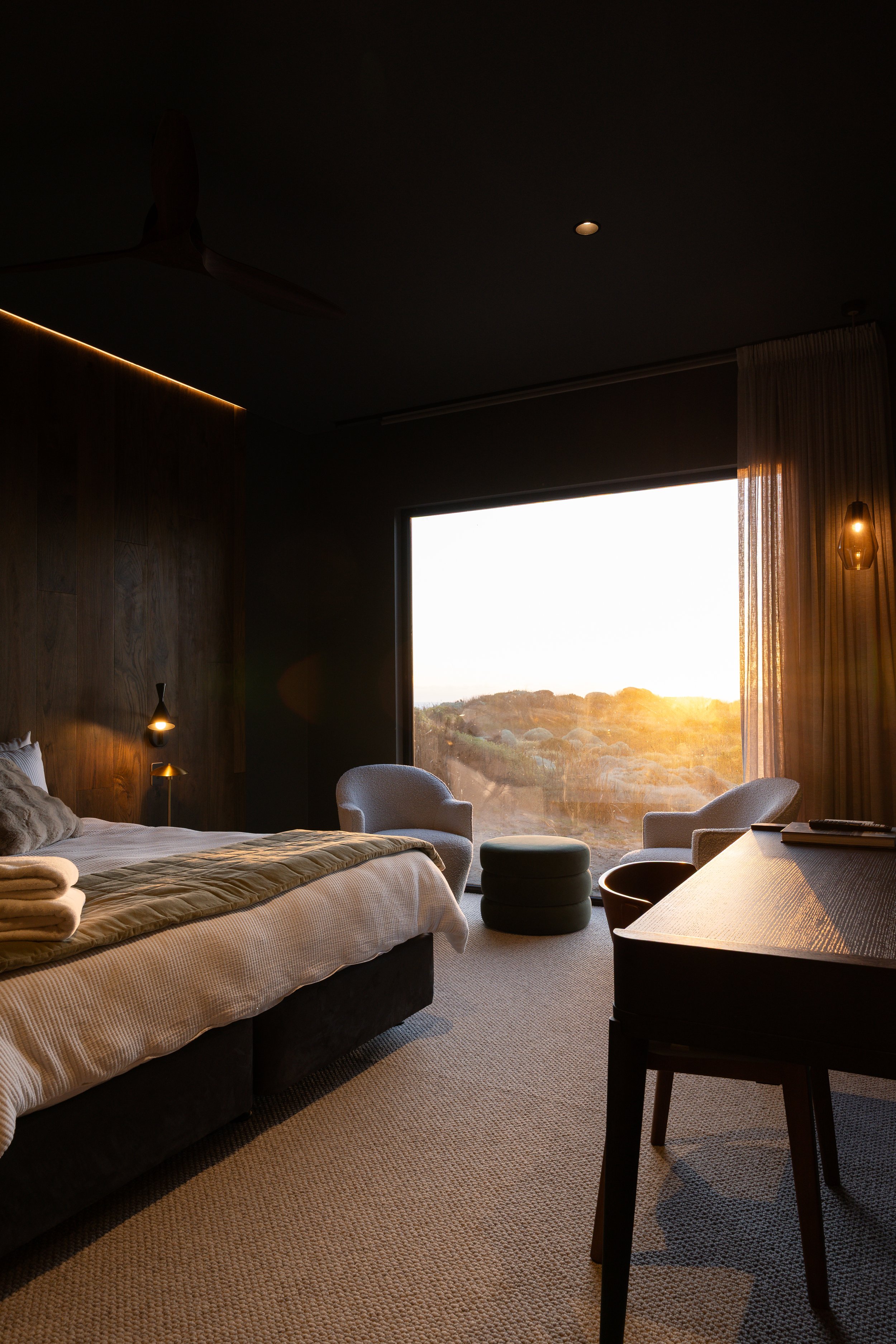 A modern hotel room with a large window showing an outdoor sunset landscape. The room has a bed with a beige cover and rolled towels, a sitting area with a white armchair and round ottoman, a desk with a chair, and warm lighting fixtures.