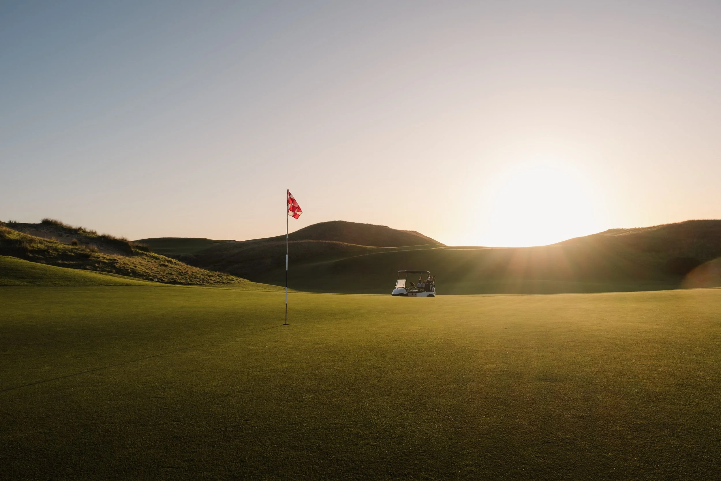 A golf course with a green, a flagstick, and a golf cart, set against rolling hills and a sunset sky.