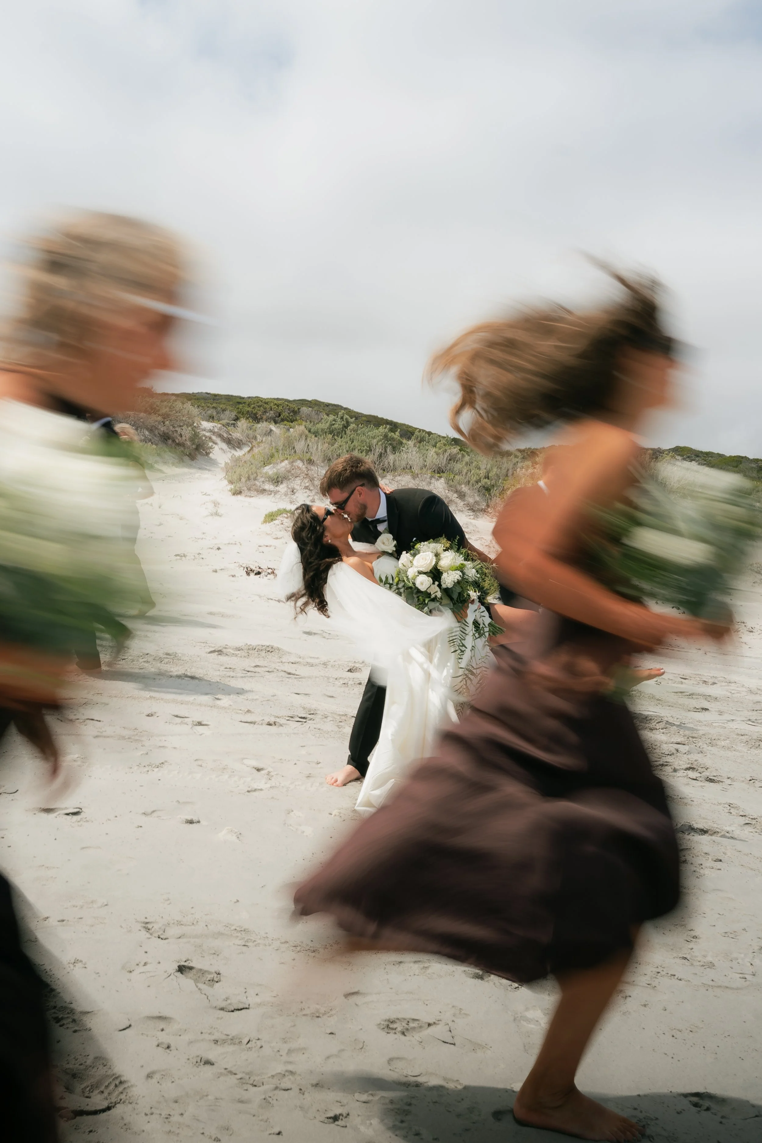 A couple in wedding attire kissing on the beach, surrounded by women walking or running past them, blurred in motion.
