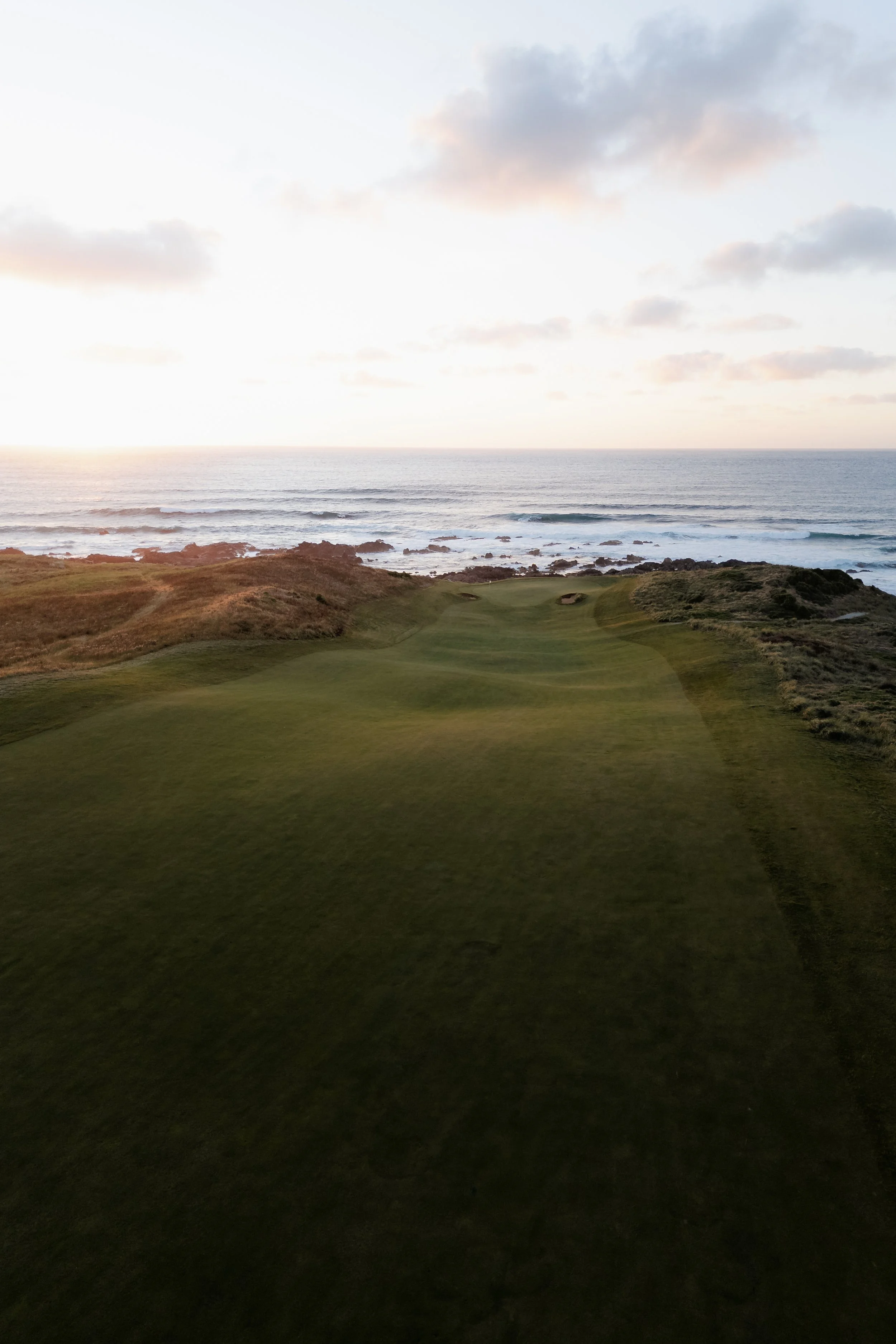 A scenic coastal landscape featuring a grassy golf course in the foreground, rocky shoreline with waves crashing against rocks, and the ocean extending to the horizon under a partly cloudy sky during sunset.