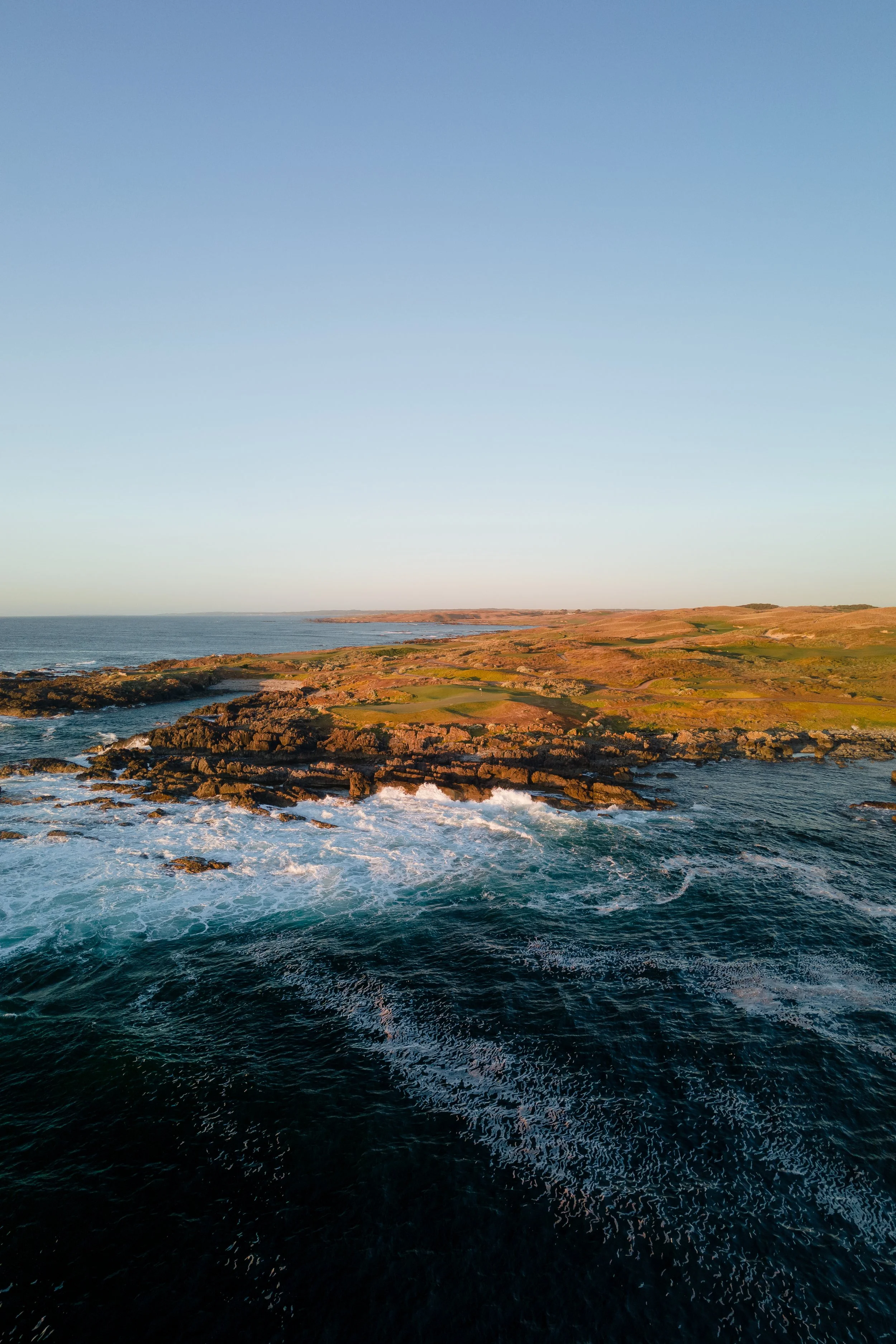 Coastal landscape with rocky shoreline, grassy hills, and calm ocean waters under a clear blue sky.
