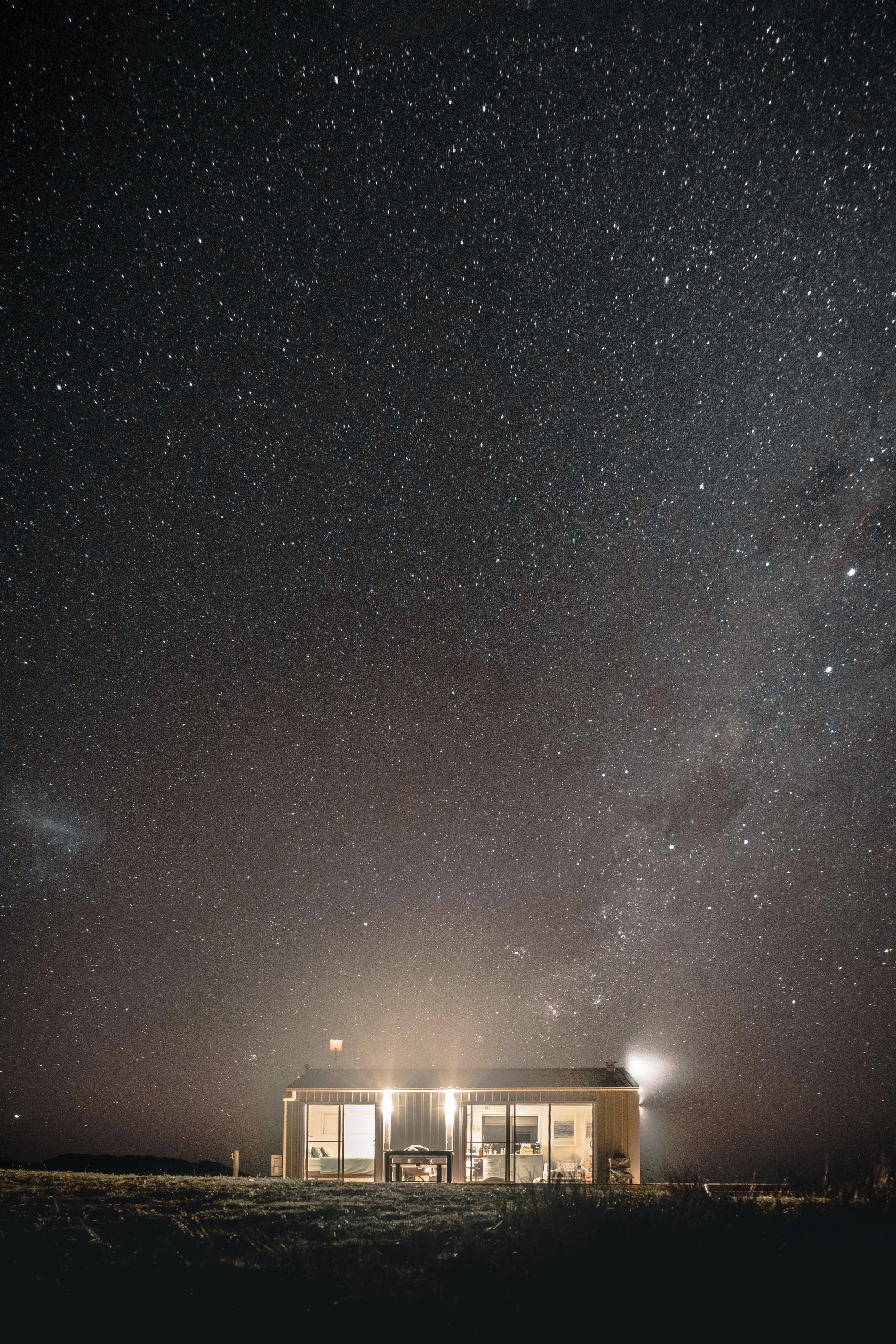 A house with bright interior lights, large windows, and a dining table visible inside, set against a backdrop of a starry night sky with countless stars and a faint glow near the horizon.