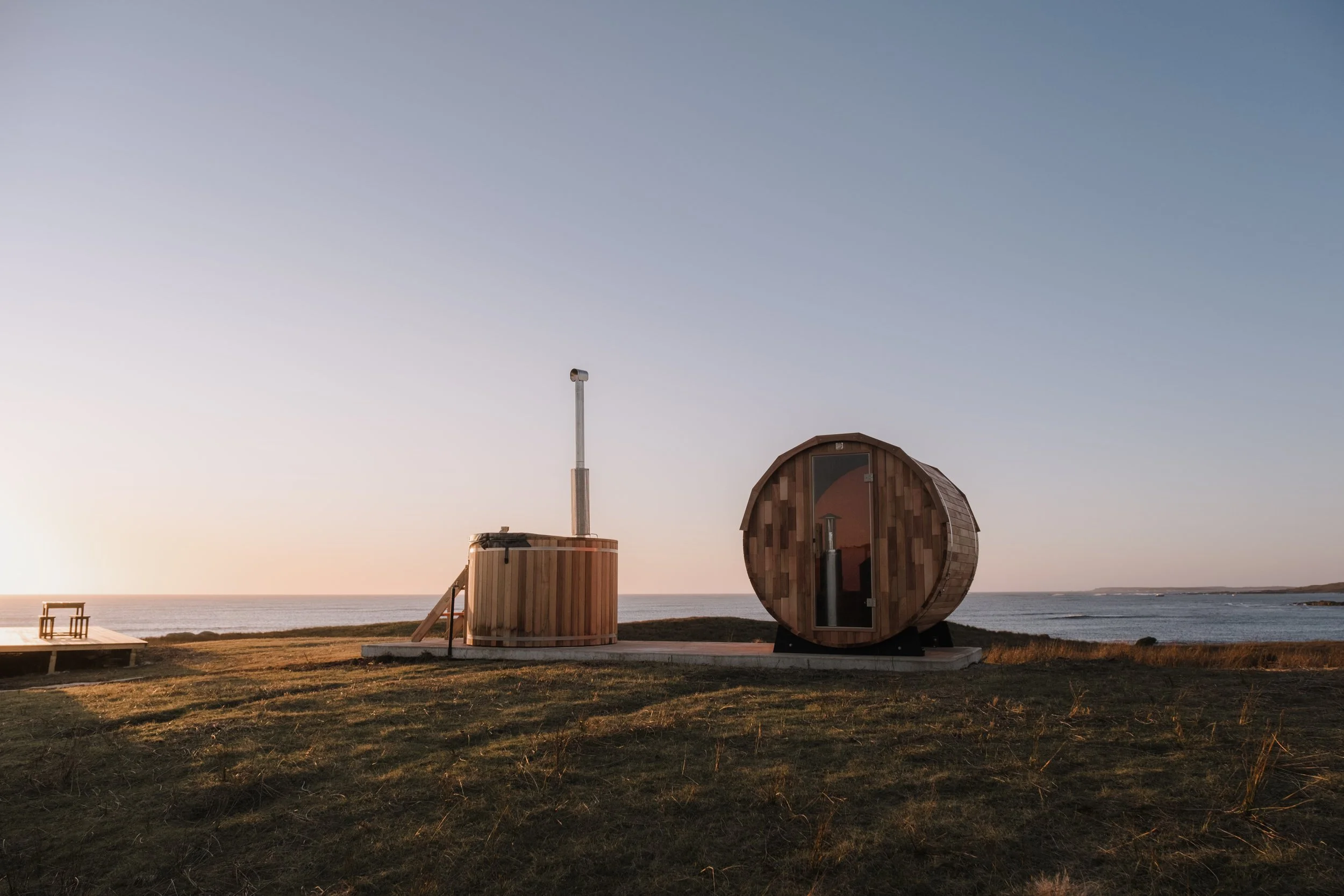 A wooden sauna with a round shape and a door, located on a grassy area near the ocean, during sunset.