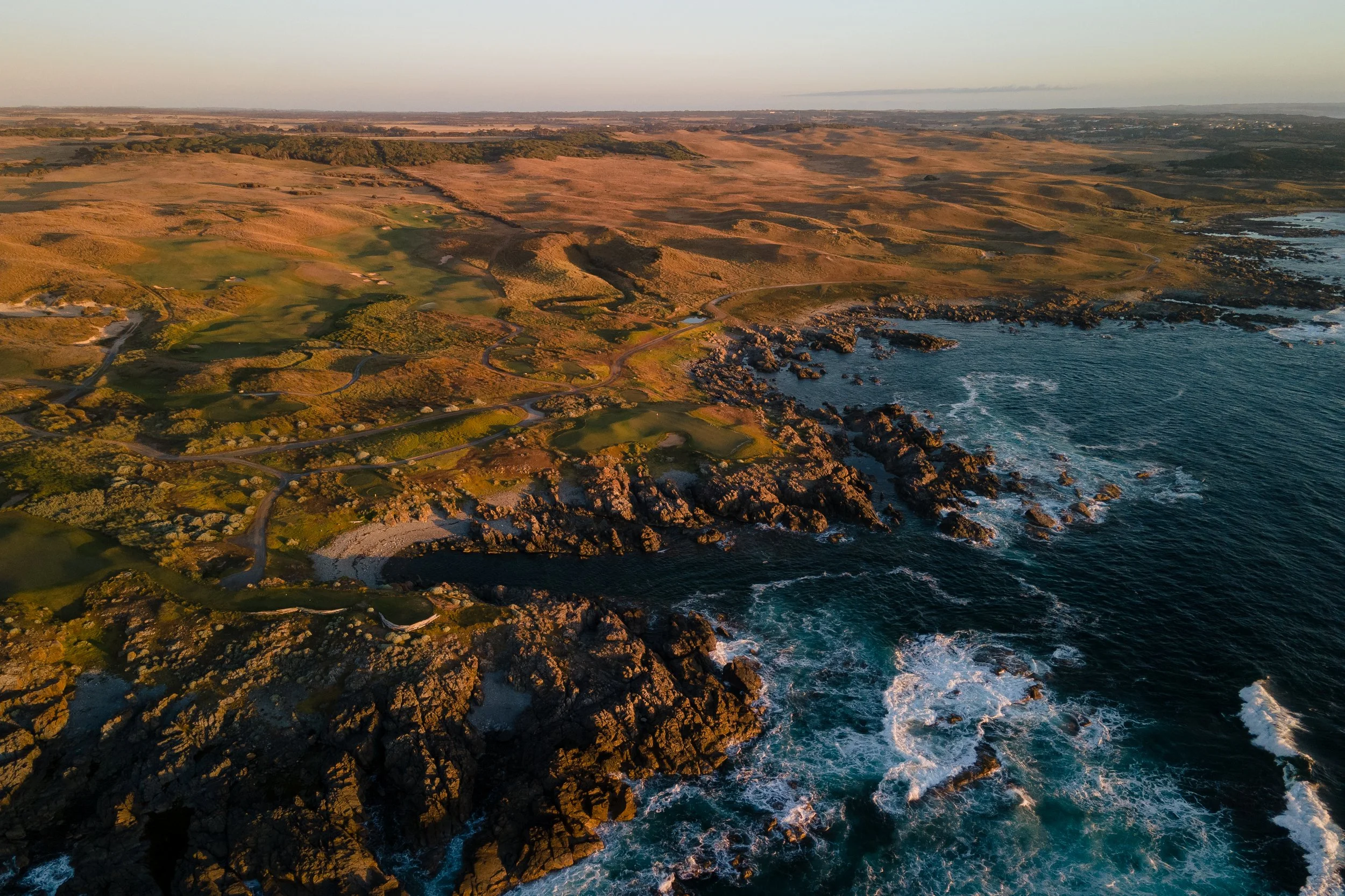 An aerial view of a coastal landscape with rocky shoreline, waves crashing, and rolling hills with sparse vegetation, during sunset.