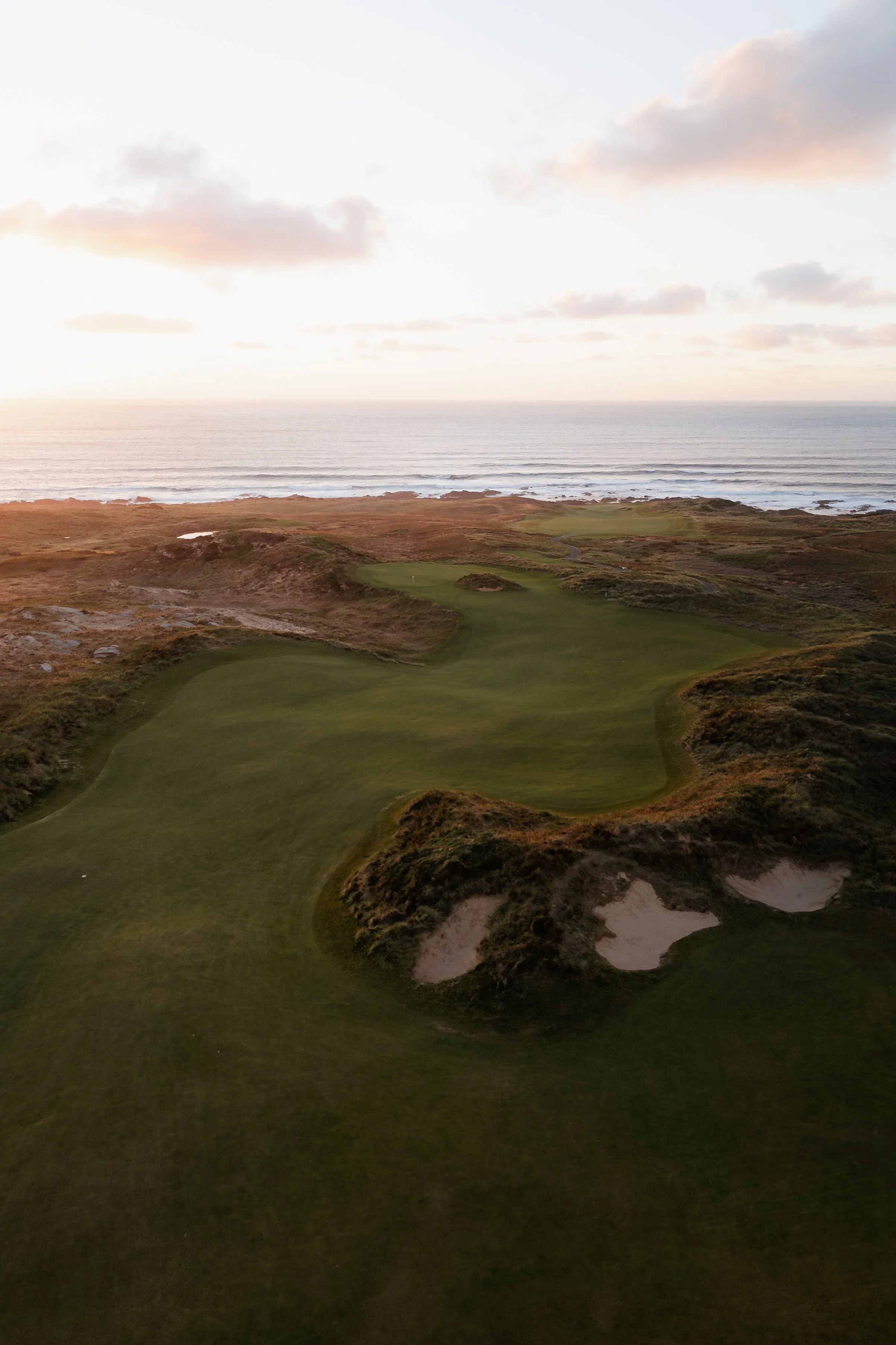 A golf course with green fairways and sand bunkers, situated along a coastline with the ocean and cloudy sky in the background.