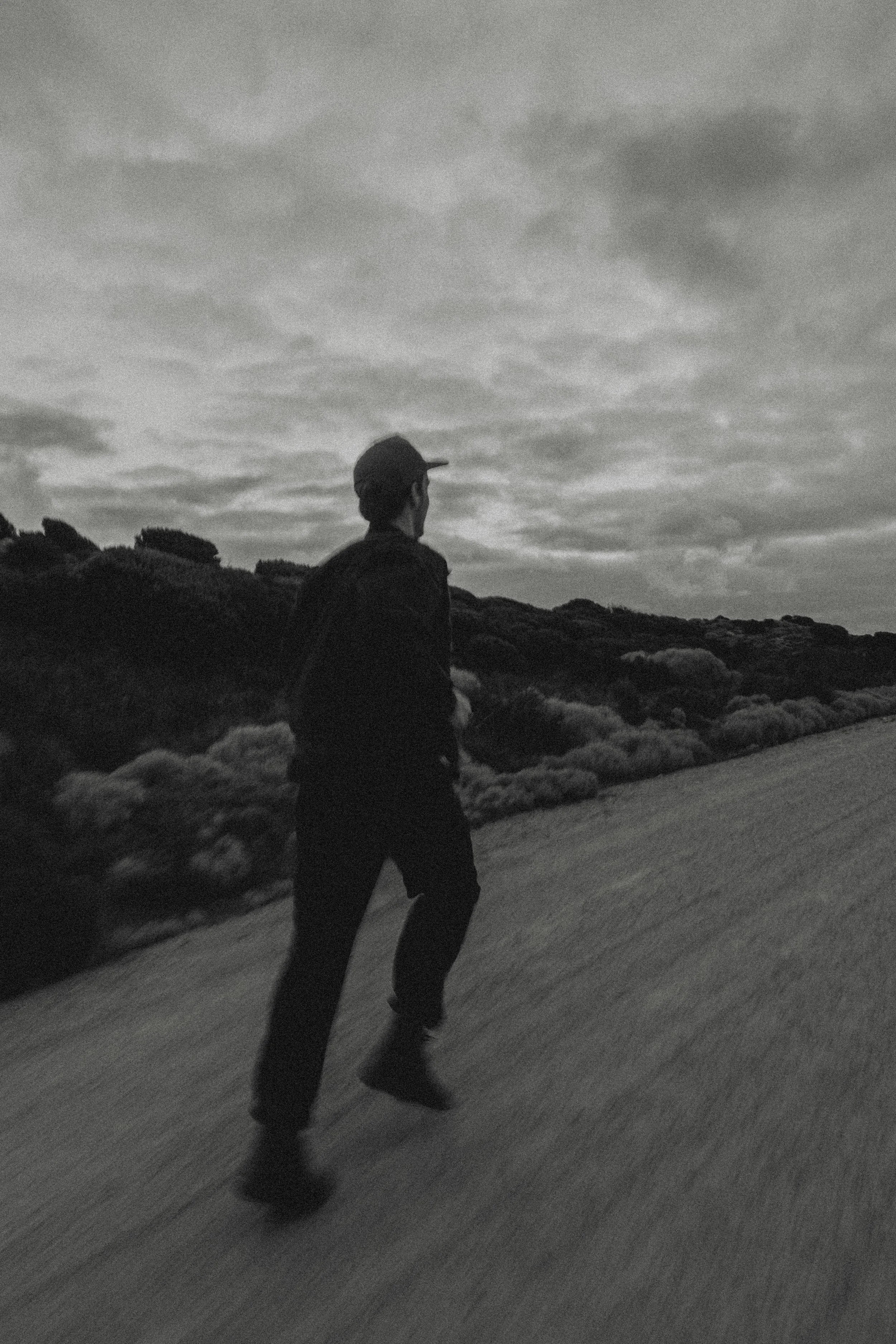 A person walking along a mountainous landscape with clouds overhead in black and white.