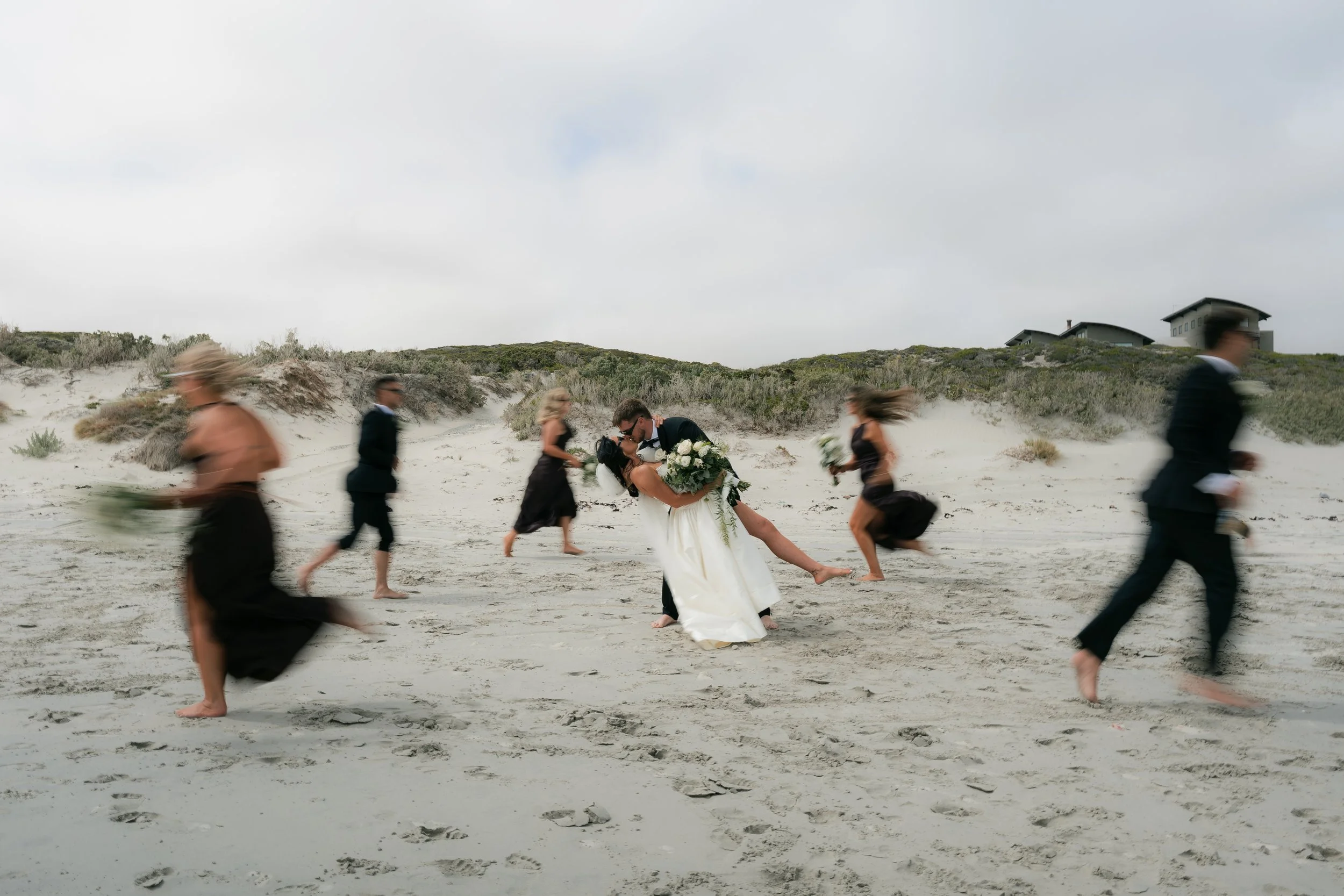 A wedding couple on a beach, with the groom kissing the bride as she holds a bouquet, surrounded by people walking on the sand with blurred motion, and houses on dunes in the background under cloudy sky.