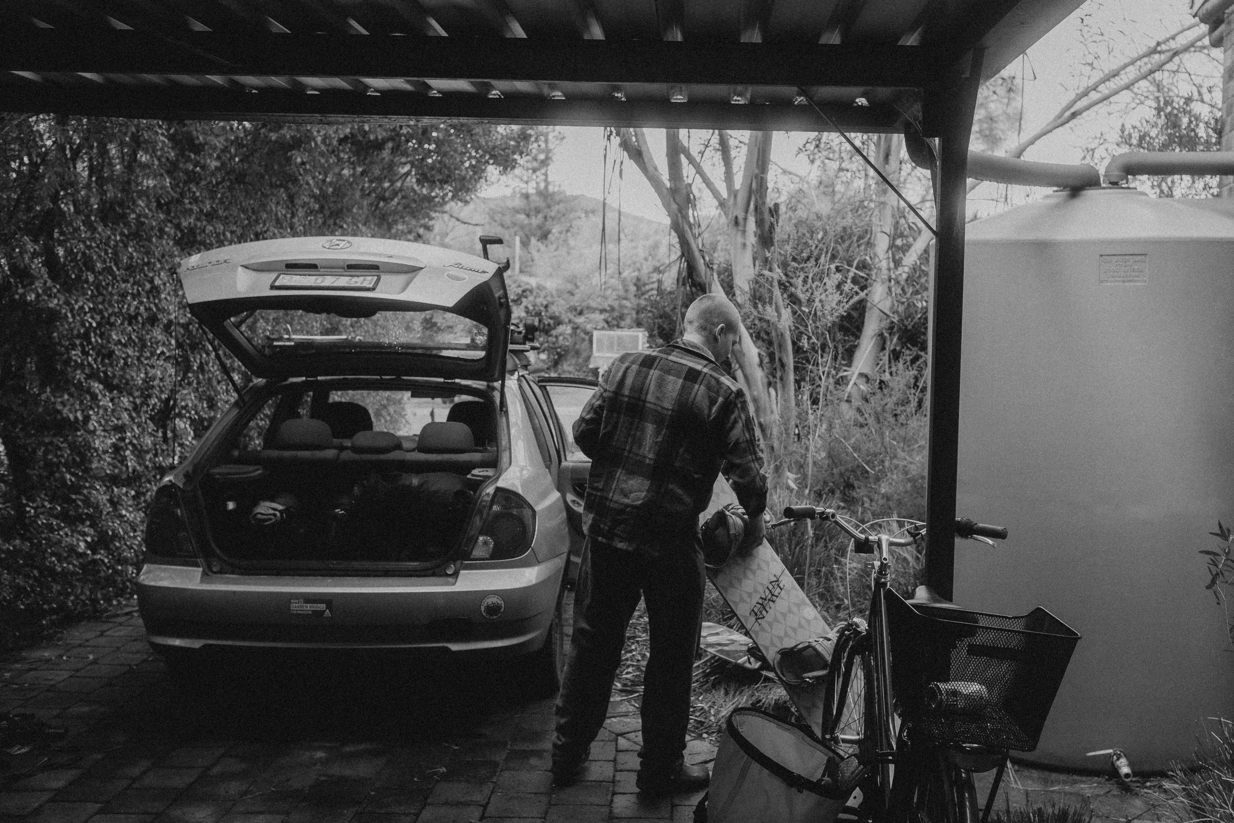 A person wearing plaid shirt standing next to a bicycle under a shelter, with a car parked behind them and open trunk, surrounded by trees and outdoor scenery.