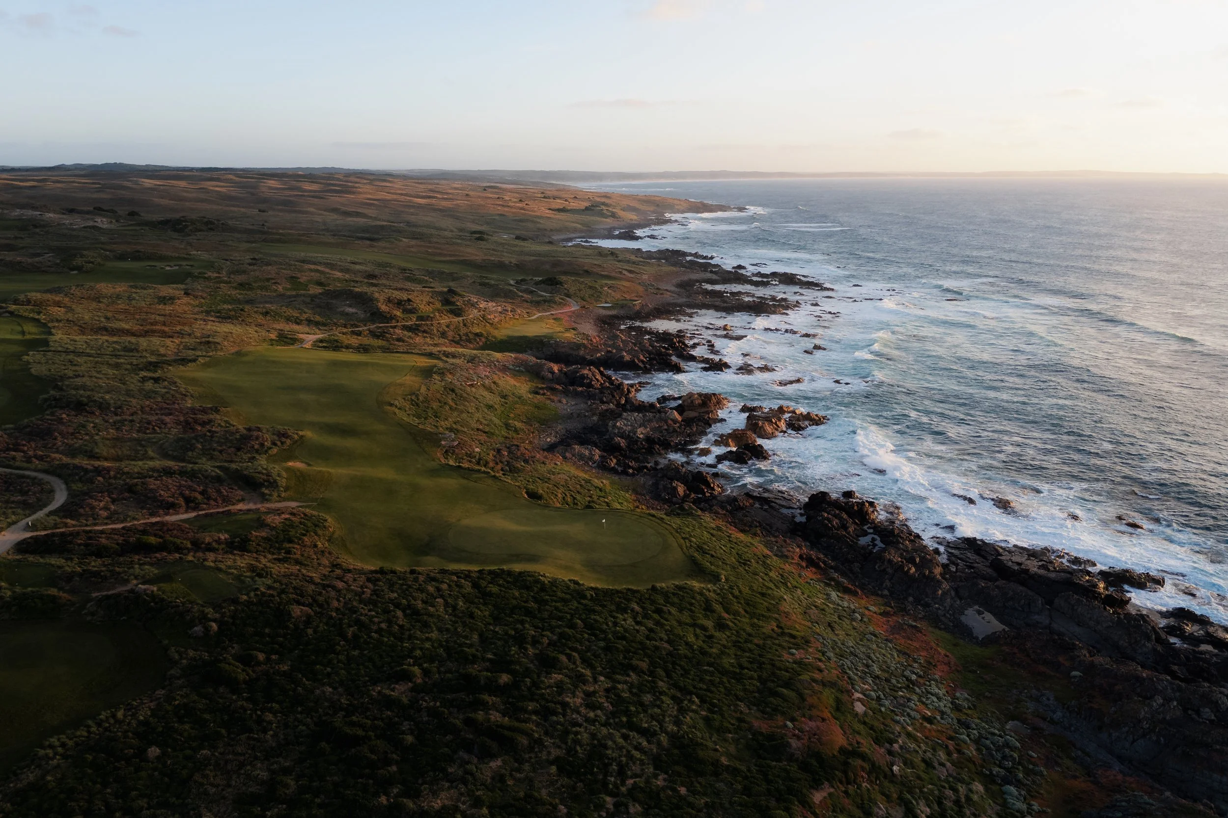 An aerial view of a rugged coastline with rocky cliffs and a golf course near the shoreline, overlooking the ocean during sunset.