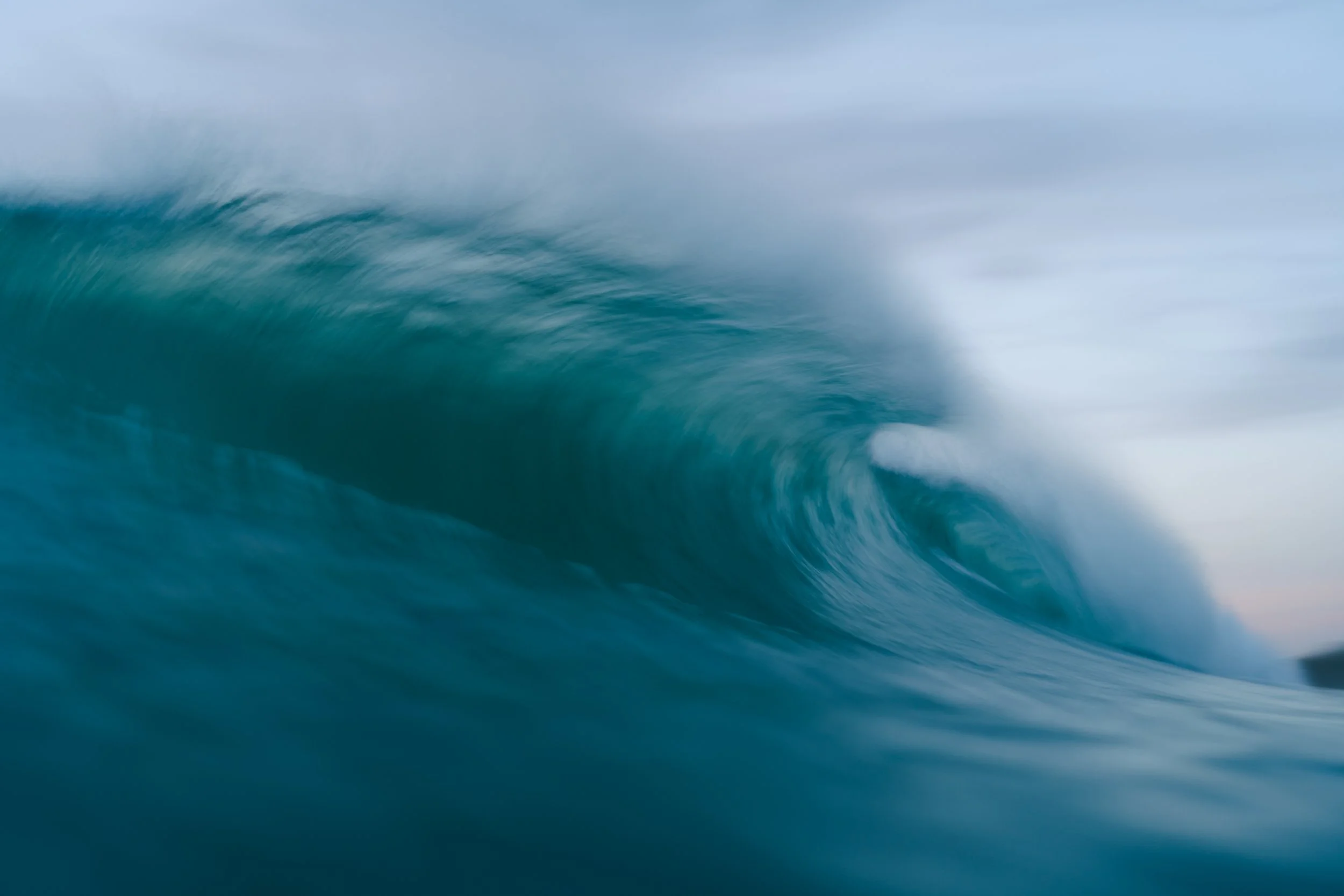 A photograph of an ocean wave with white foam and a blue-green color, captured mid-wave with a cloudy sky in the background.