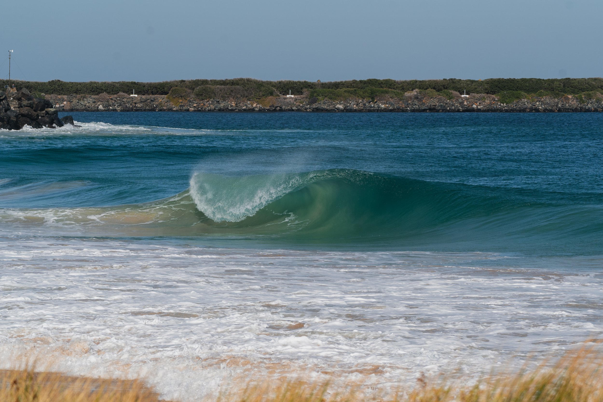 The image shows the ocean with waves crashing onto the sandy shore. There is a rocky breakwater and a strip of green vegetation with bushes in the background under a clear sky.