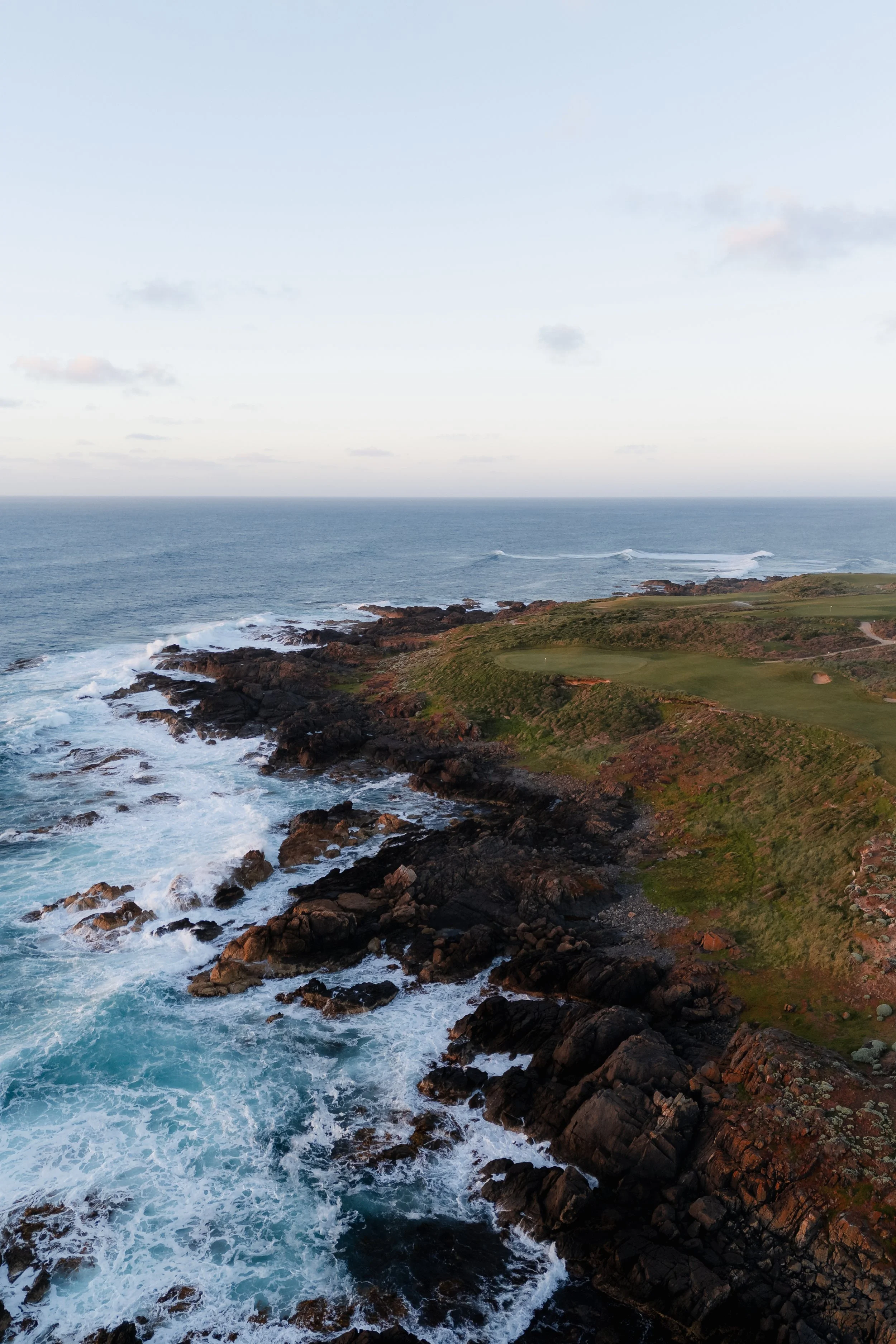 Rocky coastline with waves crashing against rocks and green grassy land in the distance under a partly cloudy sky.