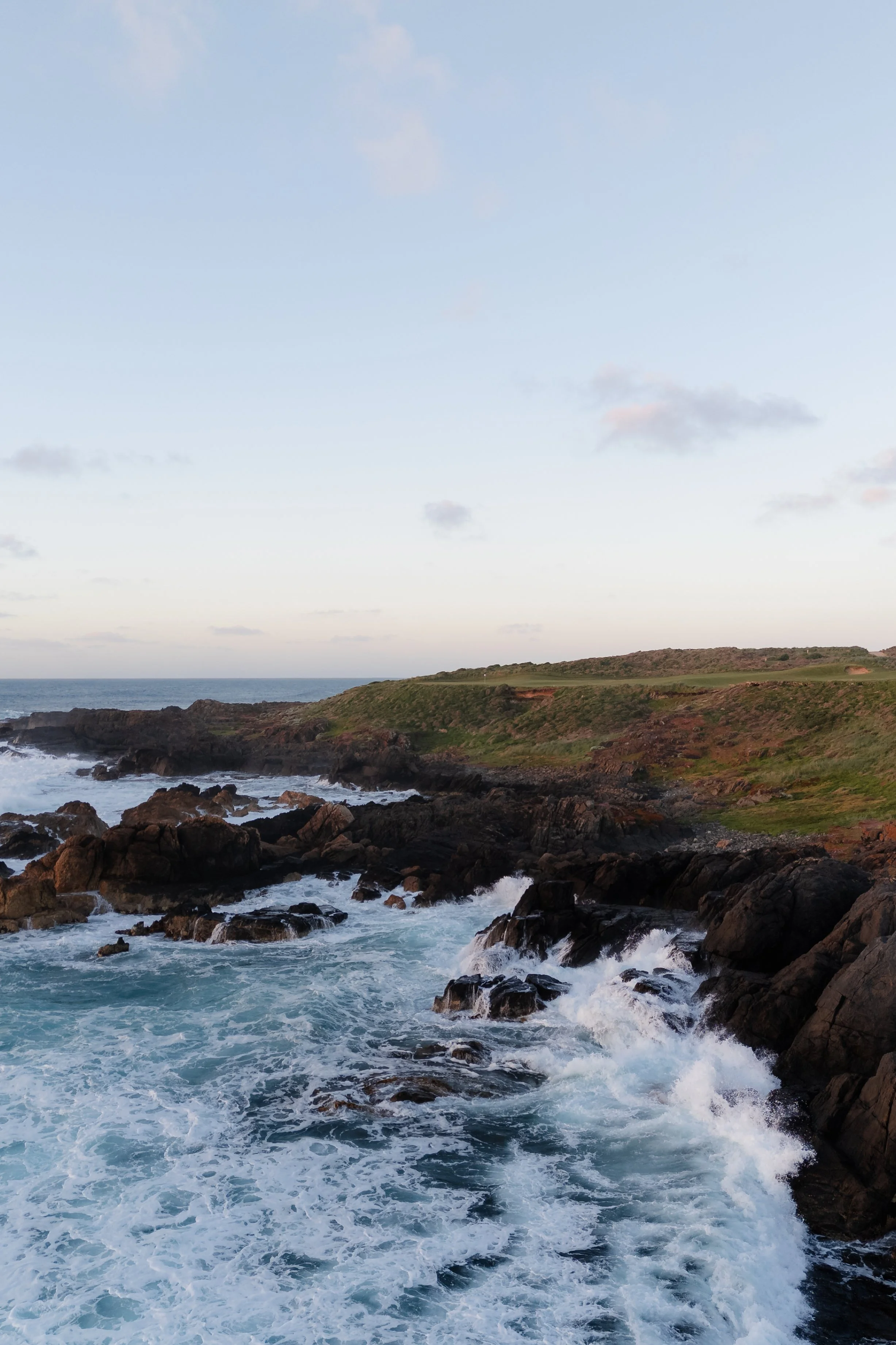 Coastal landscape with rocky shoreline, waves crashing against rocks, grassy hill, and a partly cloudy sky.