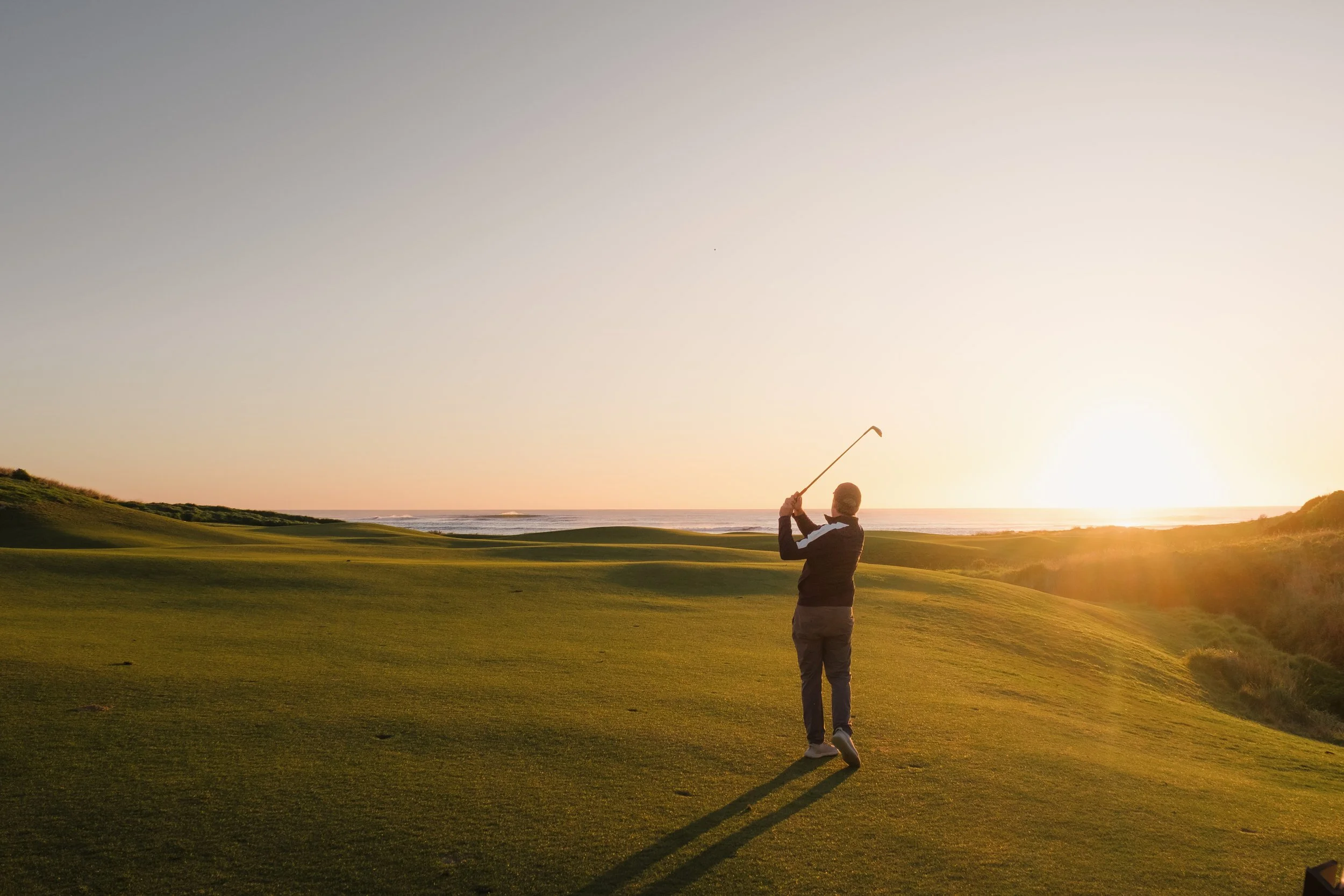 A person playing golf on a course at sunset, swinging a club with the ocean in the background.