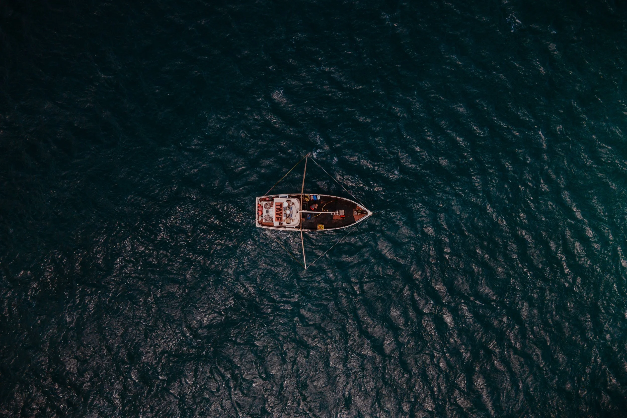 Aerial view of a sailboat in dark blue ocean water with small waves, seen from above.