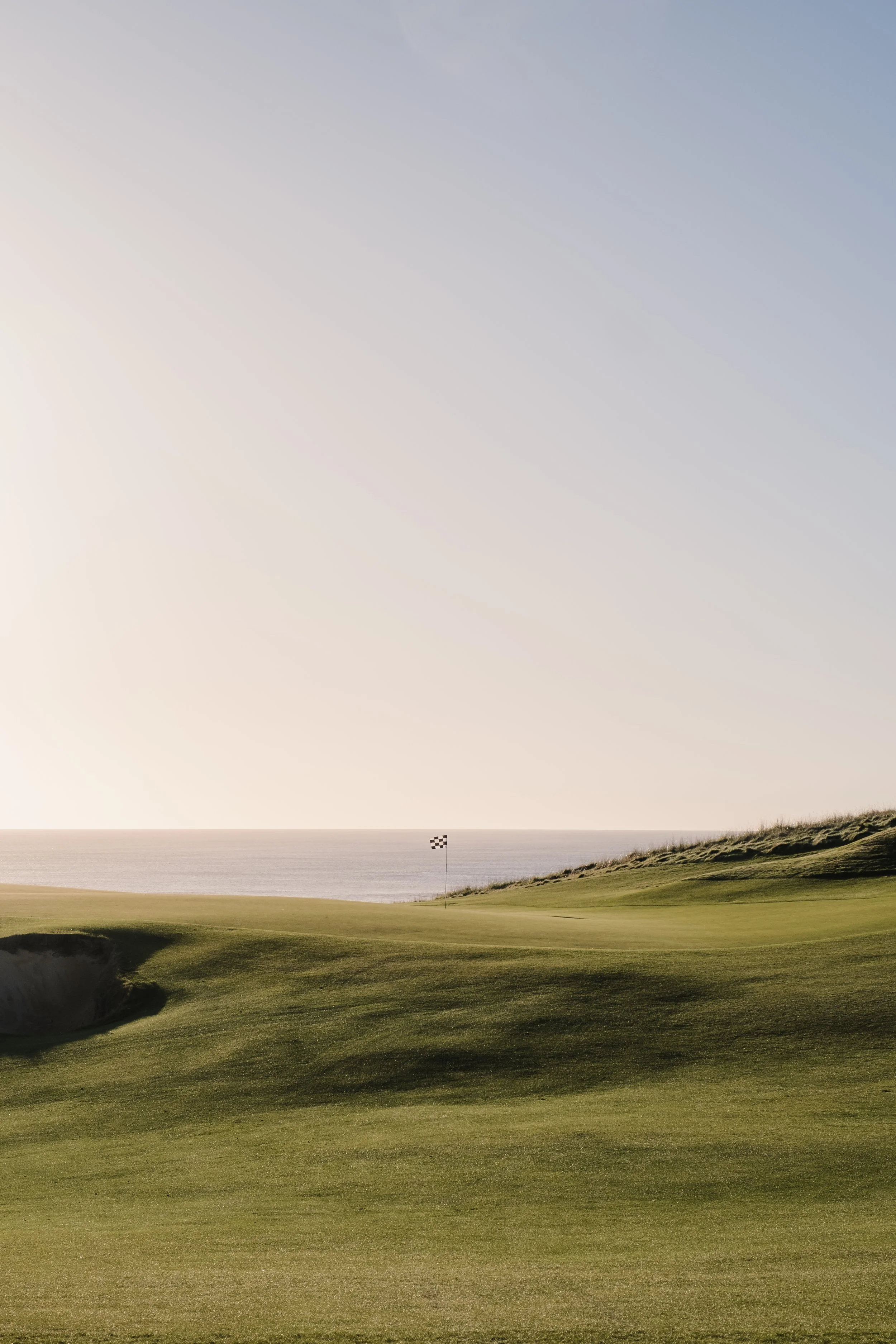 A golf course with a green and a flag sticker near the water, overlooking a large body of water and distant land under a clear sky.