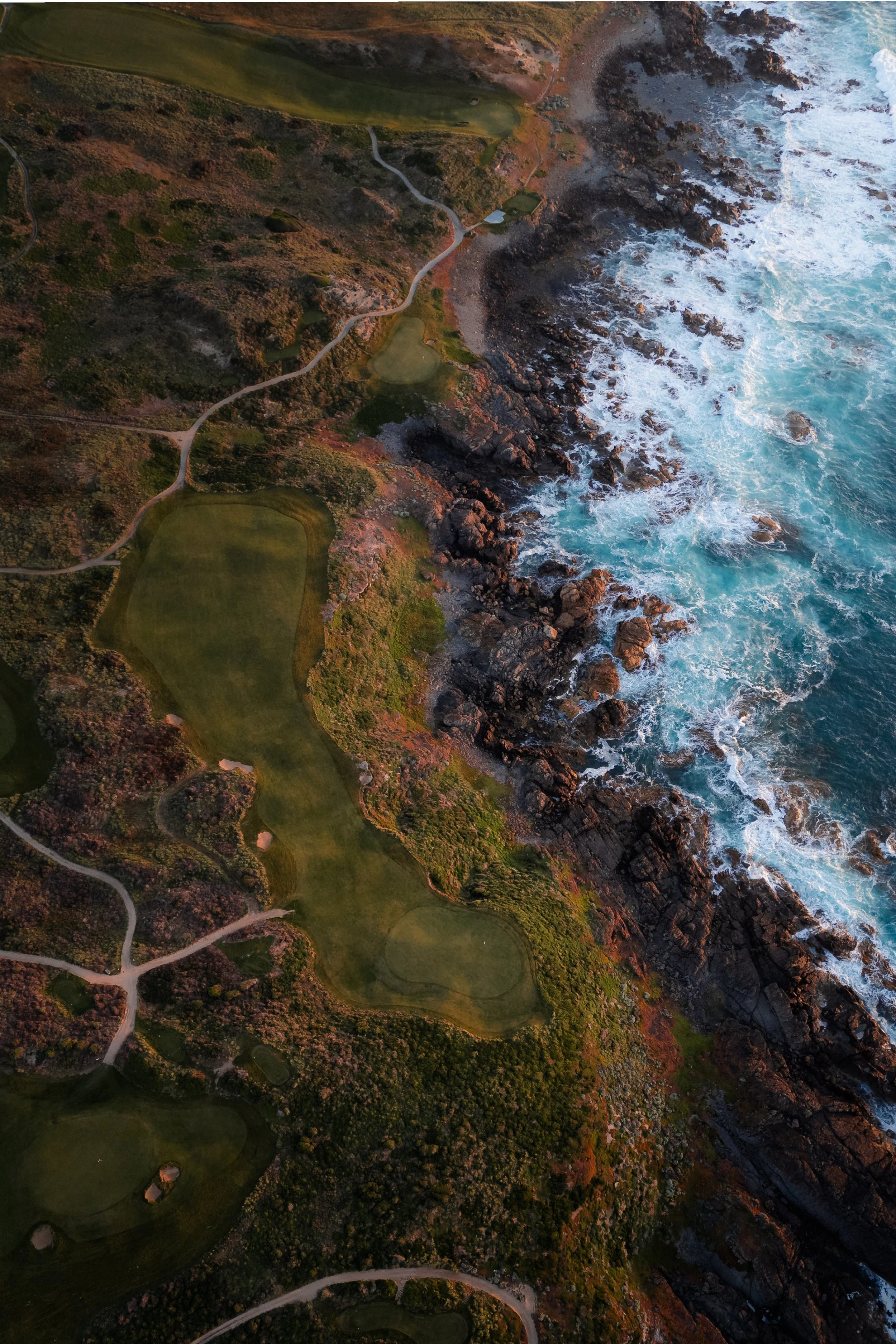 Aerial view of a coastal golf course with green fairways, winding paths, and rocky shoreline with waves crashing onto the rocks.