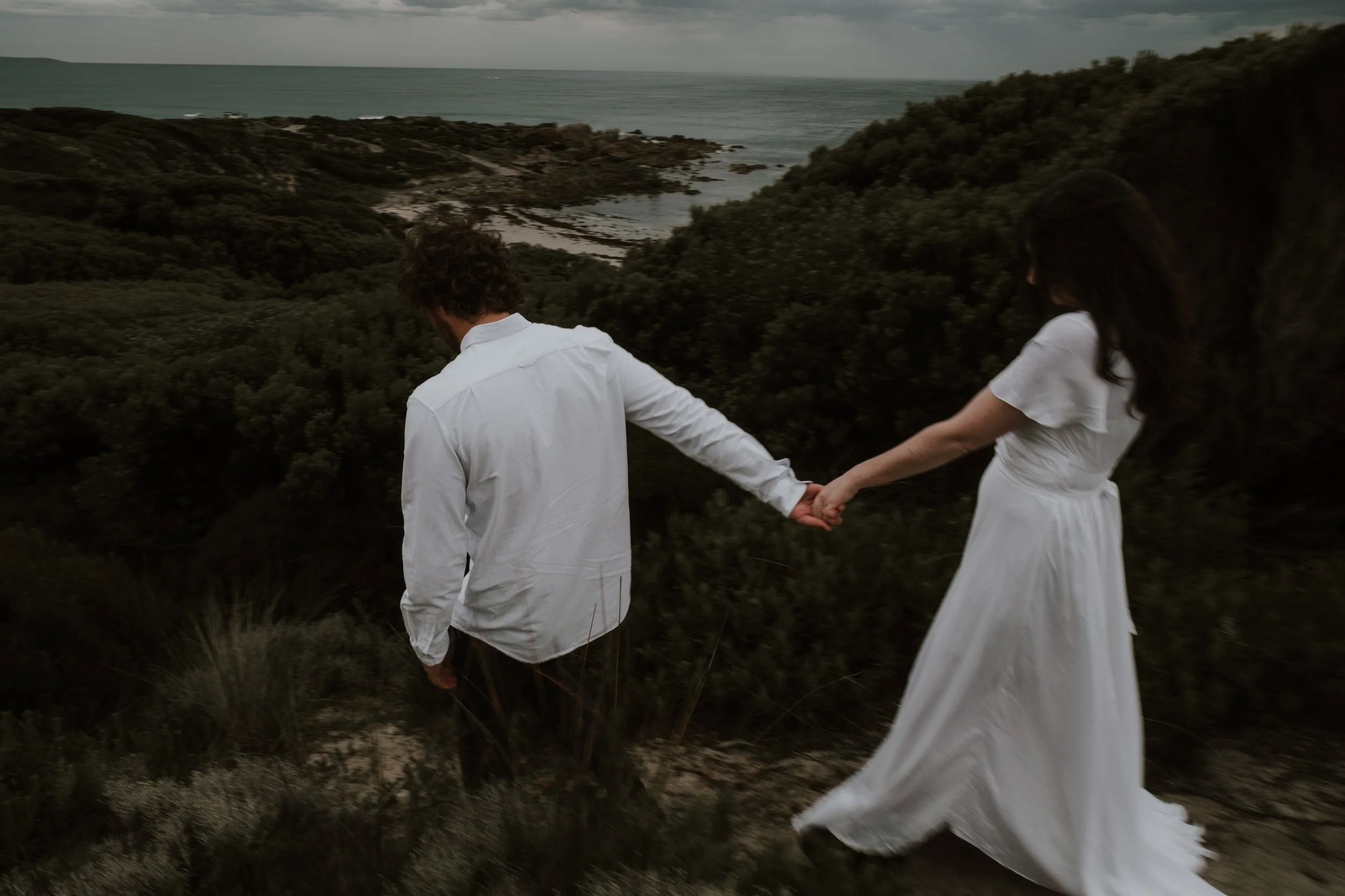 A man and a woman holding hands walking through a grassy, bushy landscape near the coast, with the ocean and cloudy sky in the background.