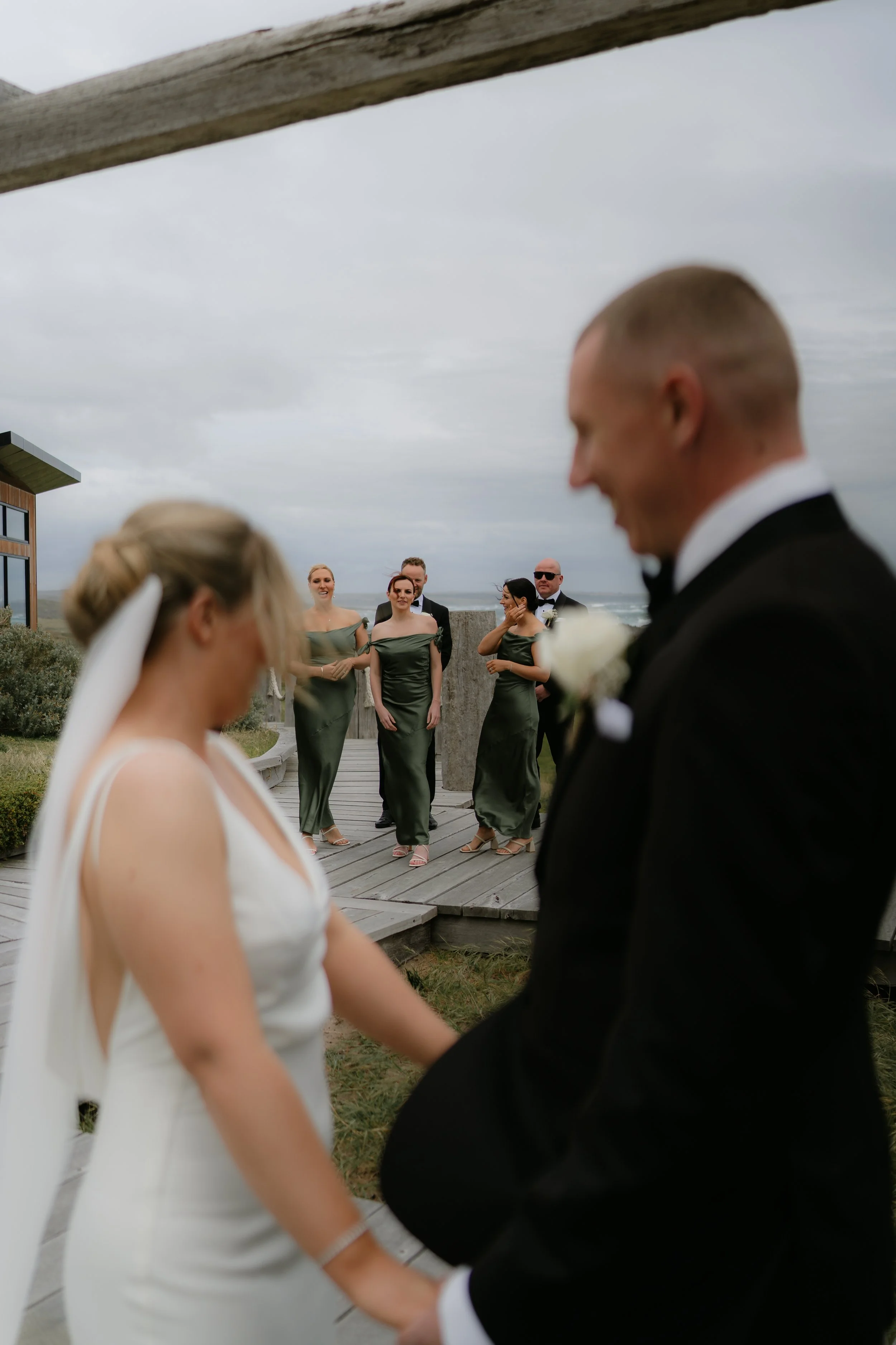 A bride and groom stand hand in hand during their outdoor wedding ceremony, with bridesmaids and groomsmen smiling in the background on a cloudy day.