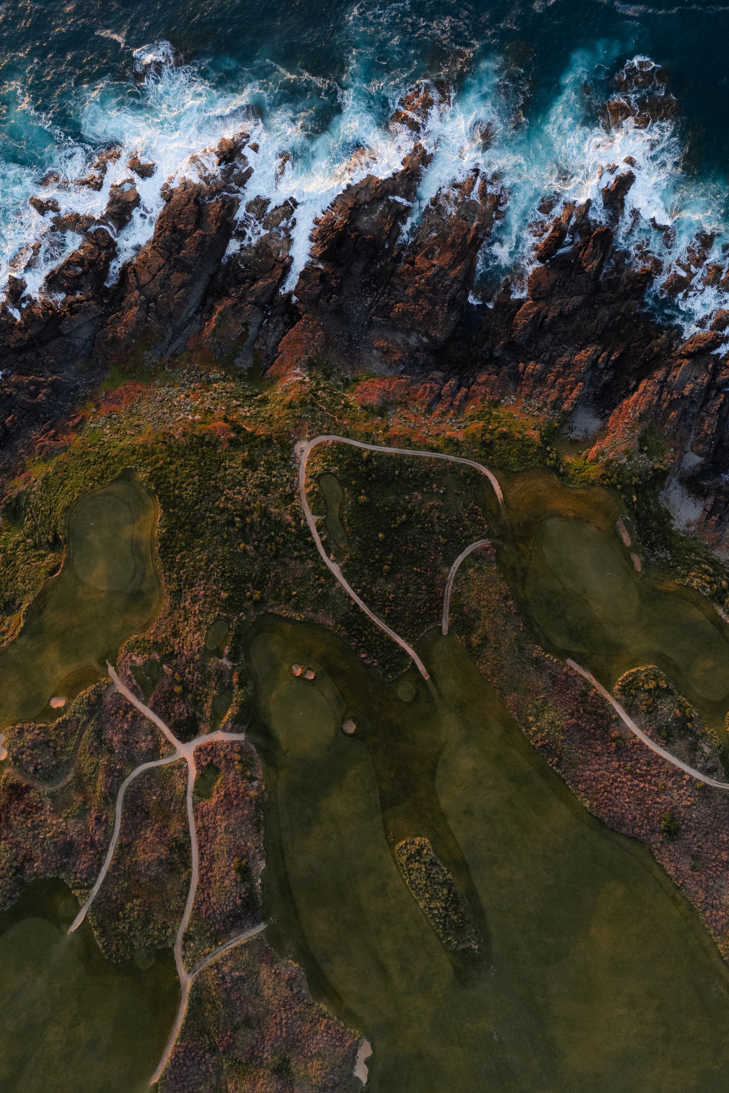 An aerial view of a rocky coastline with waves crashing against the shore, adjacent to a golf course with winding pathways and green fairways.