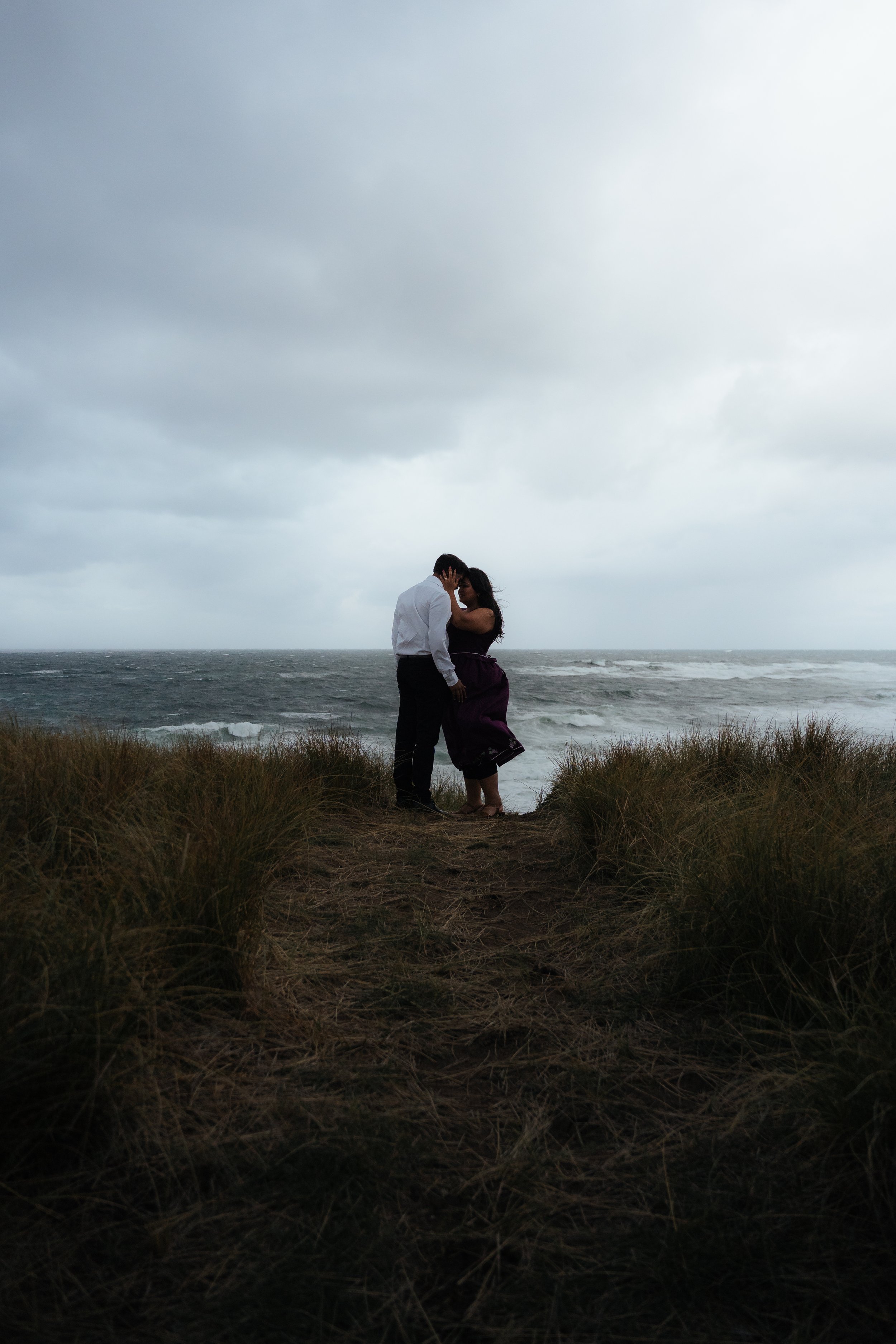 A couple stands close together on a beach, sharing an intimate moment under a cloudy sky with ocean waves in the background.