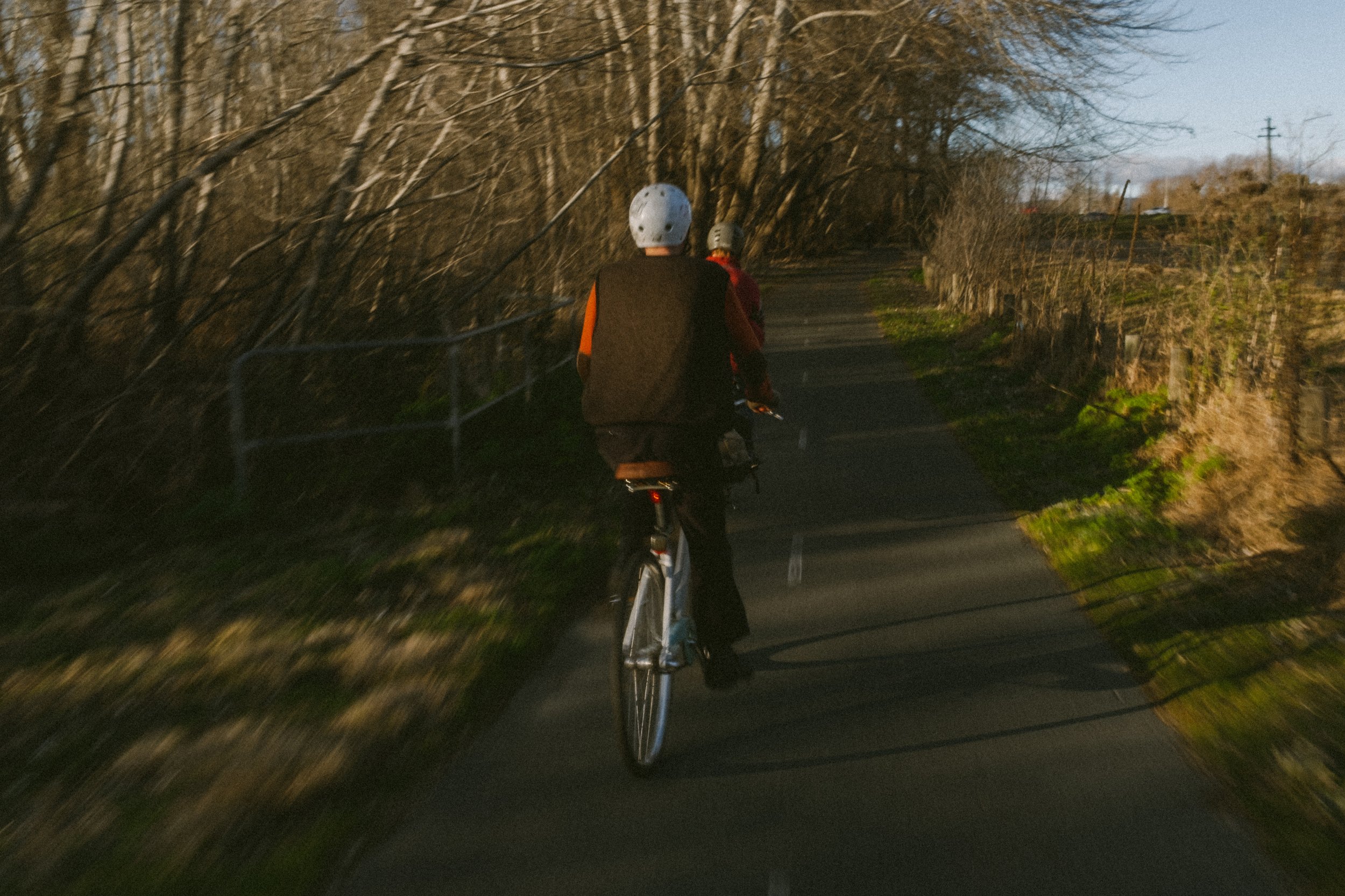 Two people riding bicycles down a narrow, tree-lined road during the daytime.