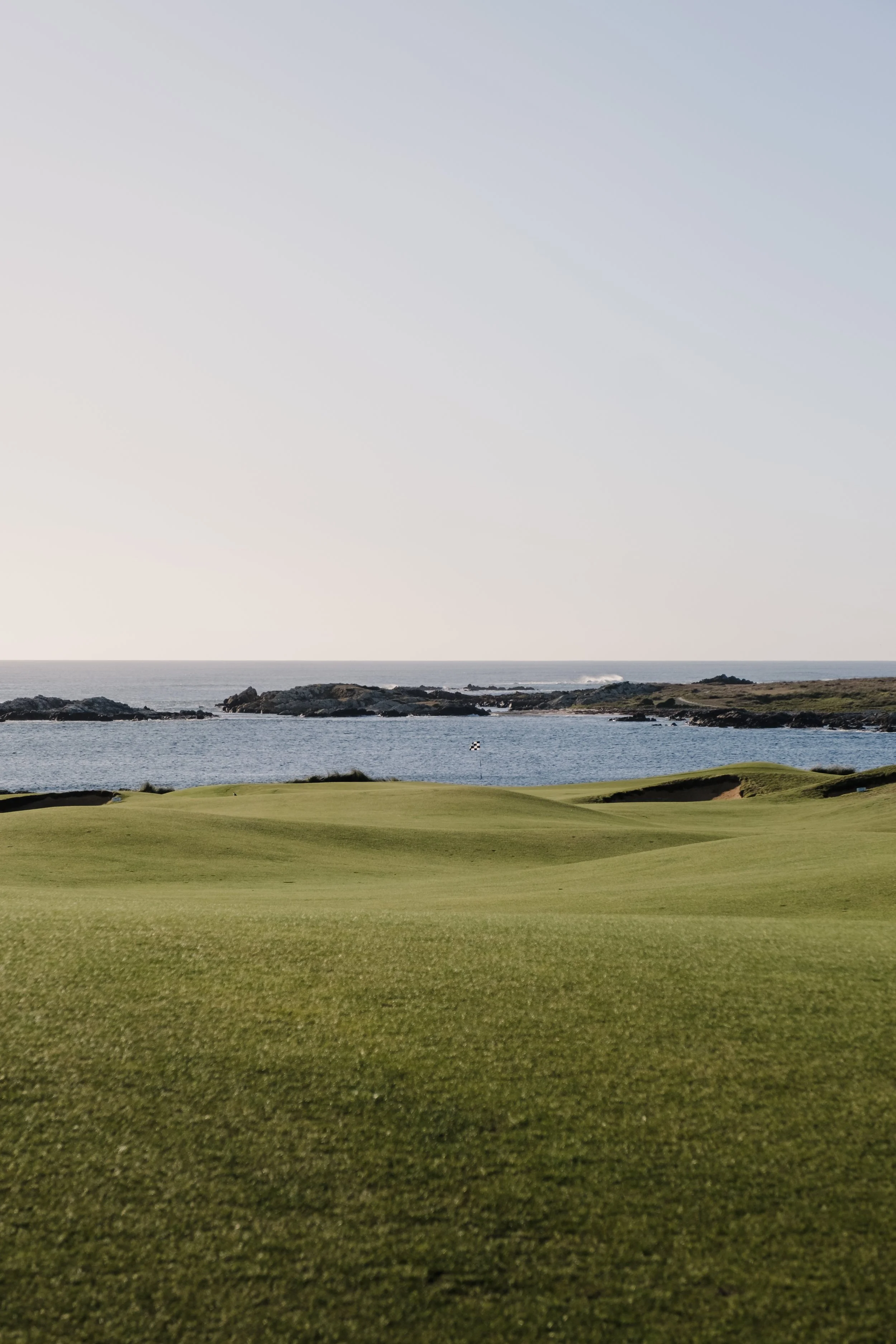 A scenic view of a golf course near the coastline, with green grass, rocky shores, and the ocean in the background under a clear sky.