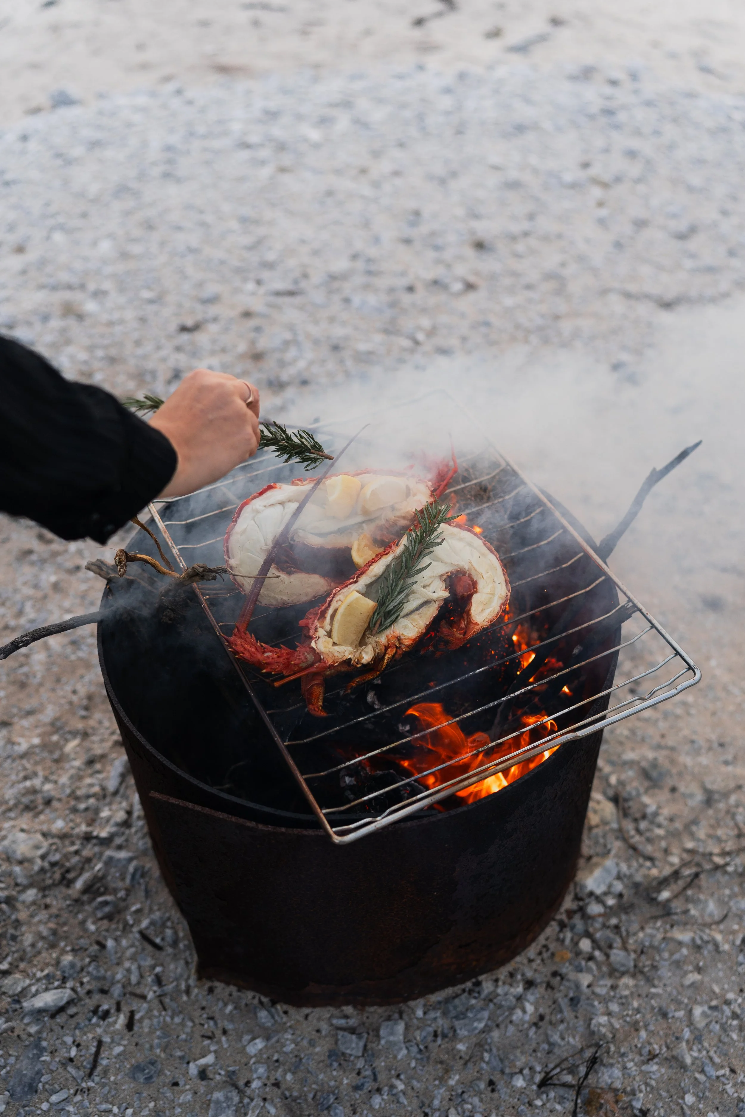 Person grilling lobster halves on an outdoor barbecue with rosemary sprigs and lemon slices.