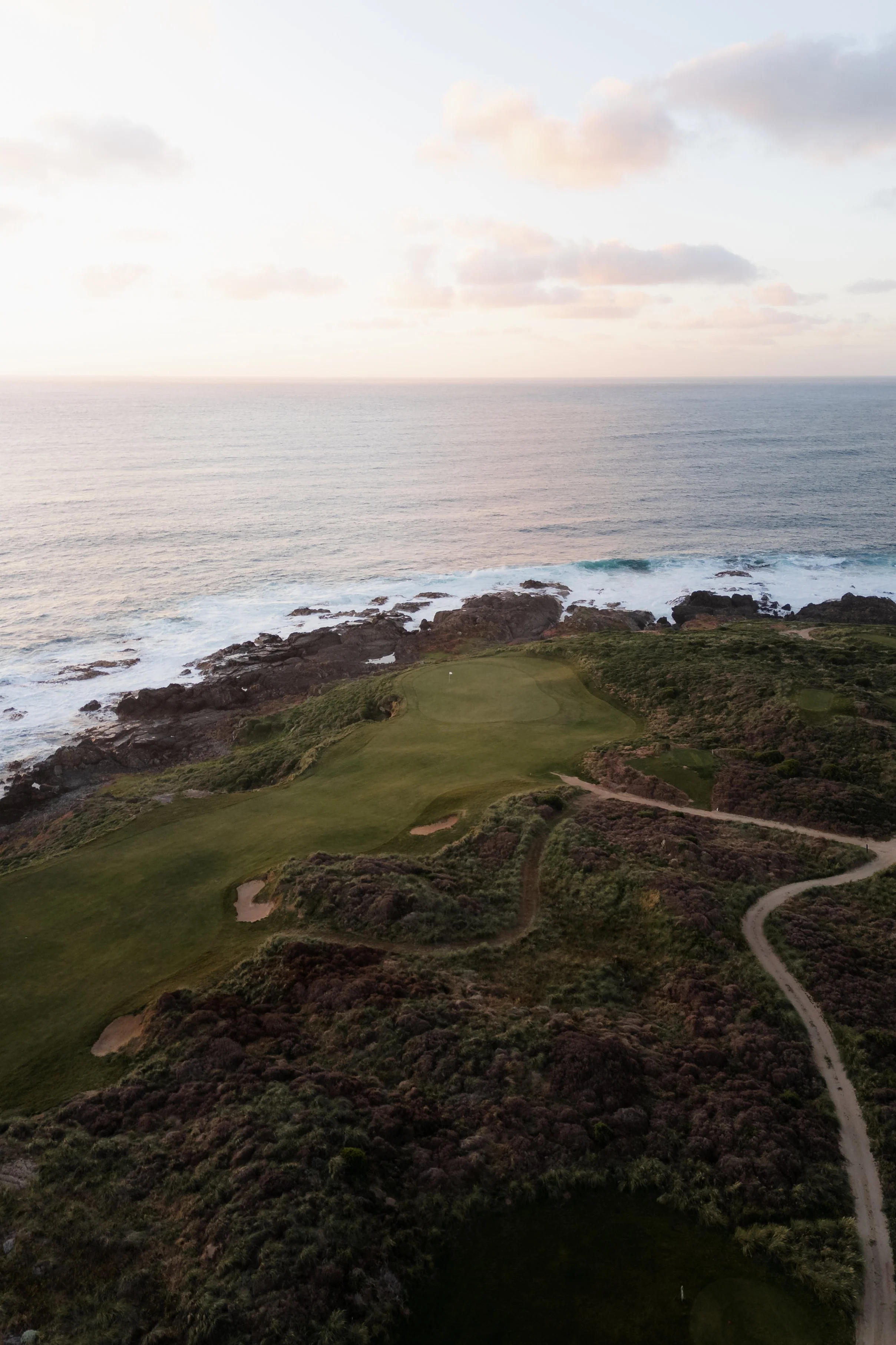 A coastal landscape with a golf course, rocky shoreline, ocean, and partly cloudy sky during sunset or sunrise.