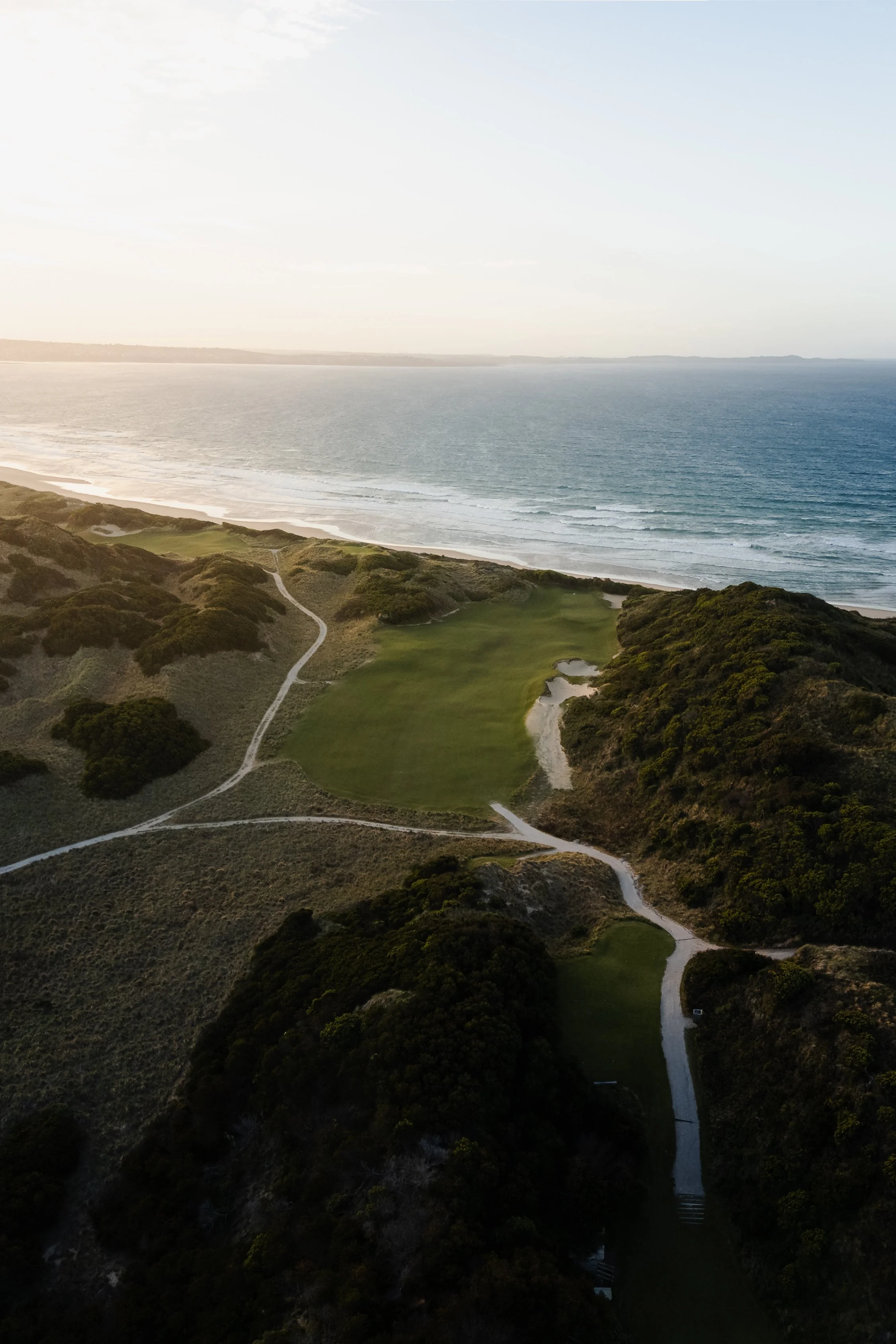 Aerial view of a coastal golf course with green fairways, surrounded by trees, overlooking the ocean with waves.