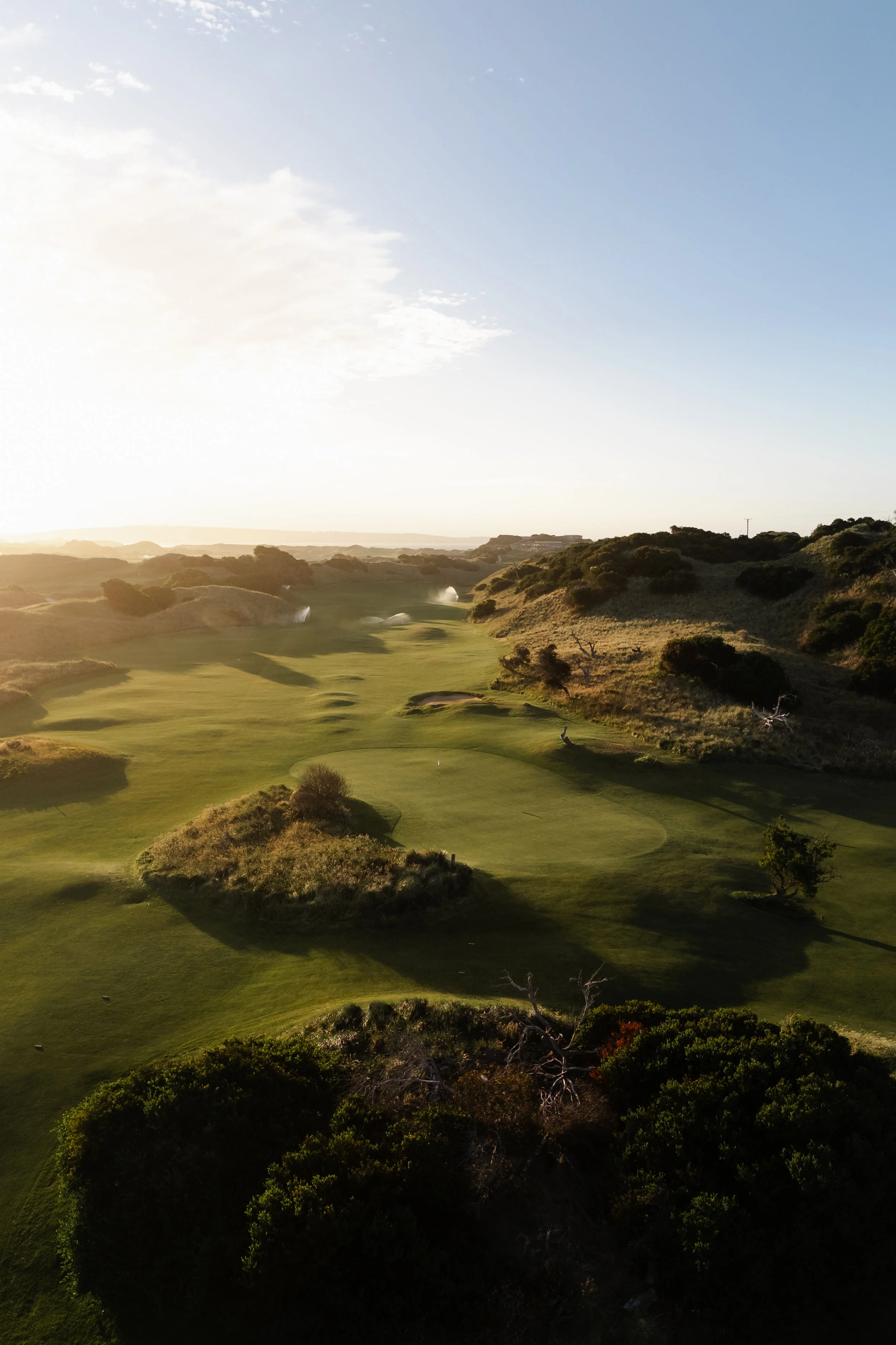 A scenic view of a golf course with rolling hills and trees, under a partly cloudy sky during sunrise or sunset.