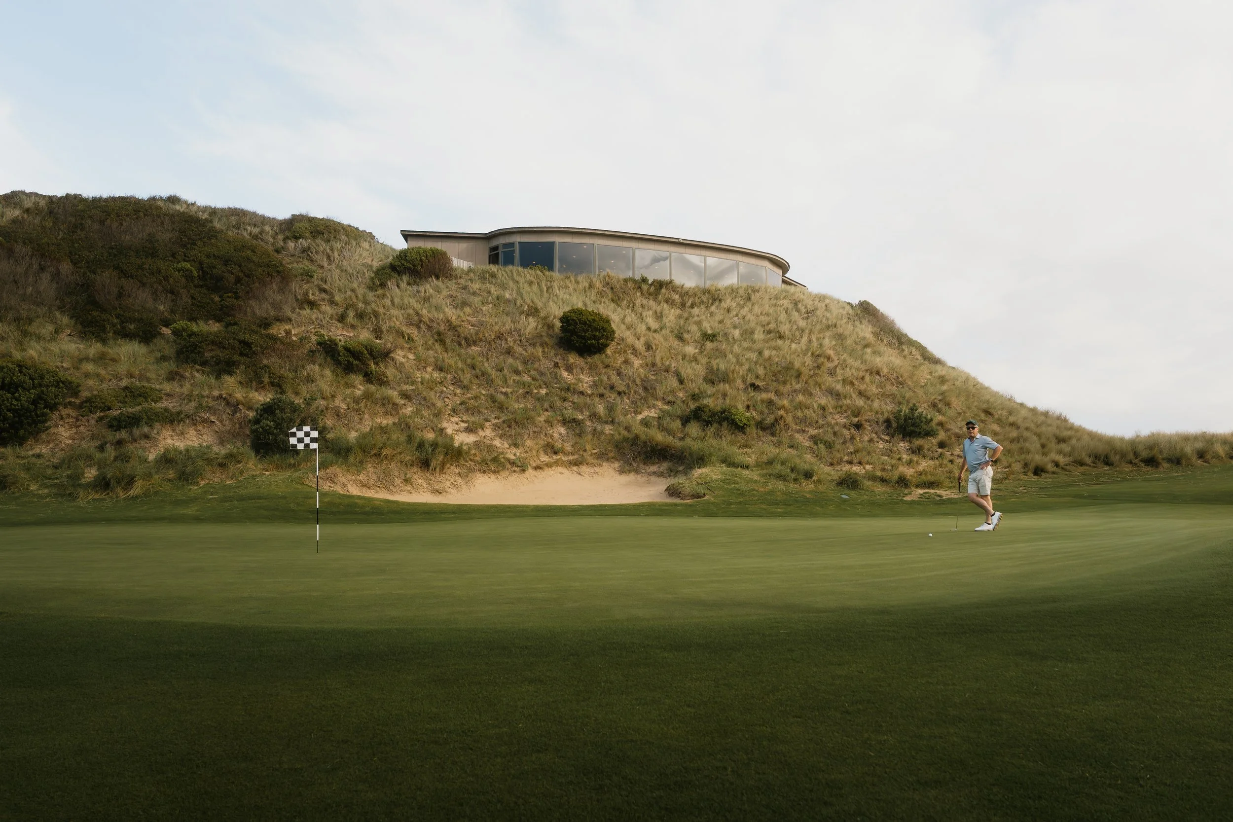 A man playing golf on a green golf course near a hillside with a modern building on top, trees, and a cloudy sky in the background.