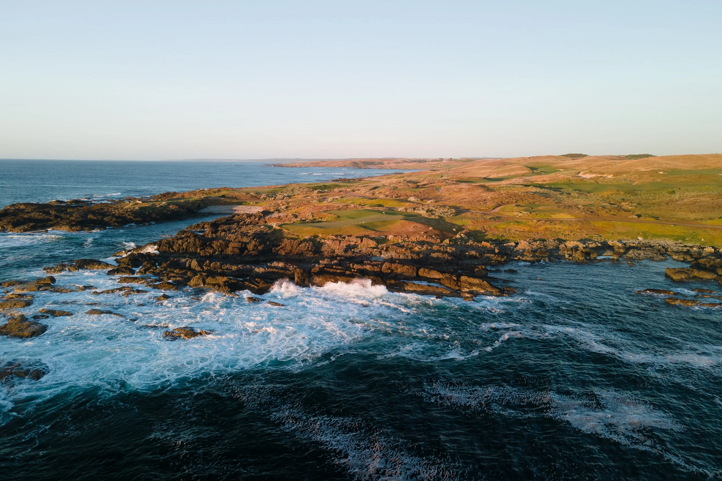 Coastal landscape with rocky shore, waves crashing, green and brown rolling hills under a clear sky.