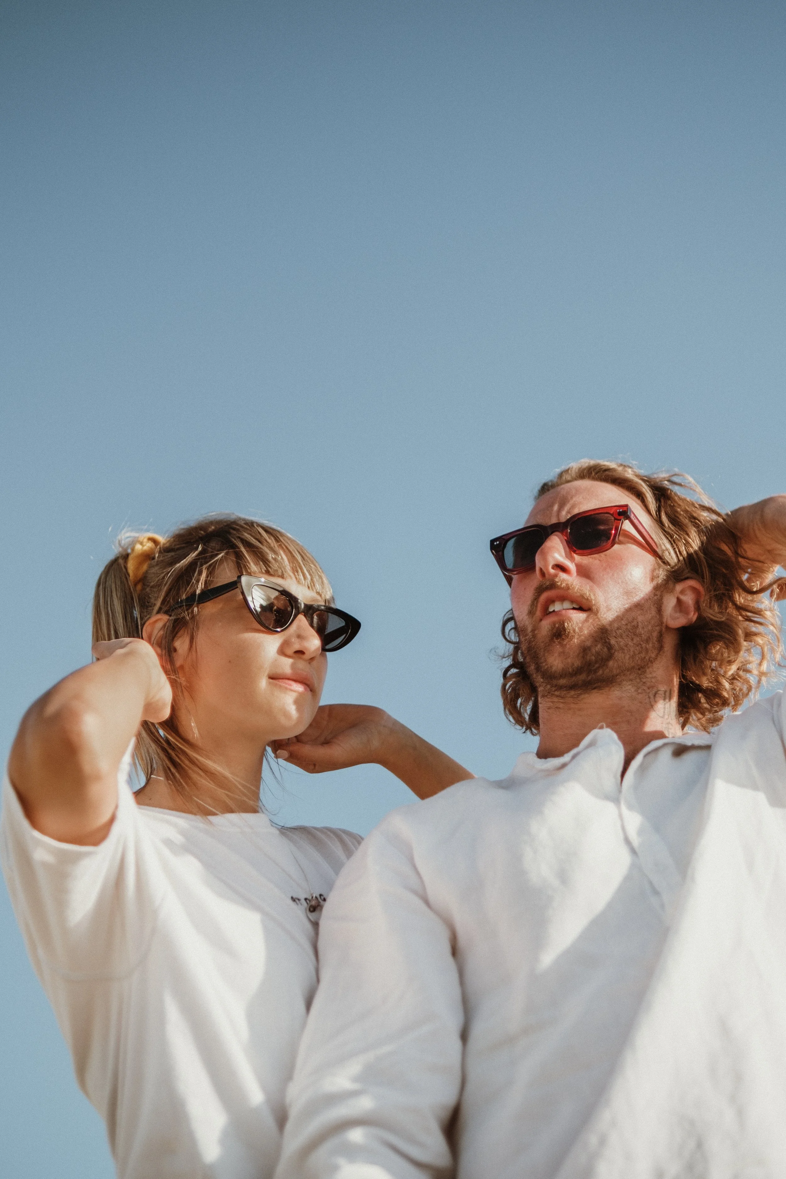 A man with long curly hair and sunglasses and a woman with straight hair and sunglasses, both wearing white shirts, standing close together outdoors against a clear blue sky.